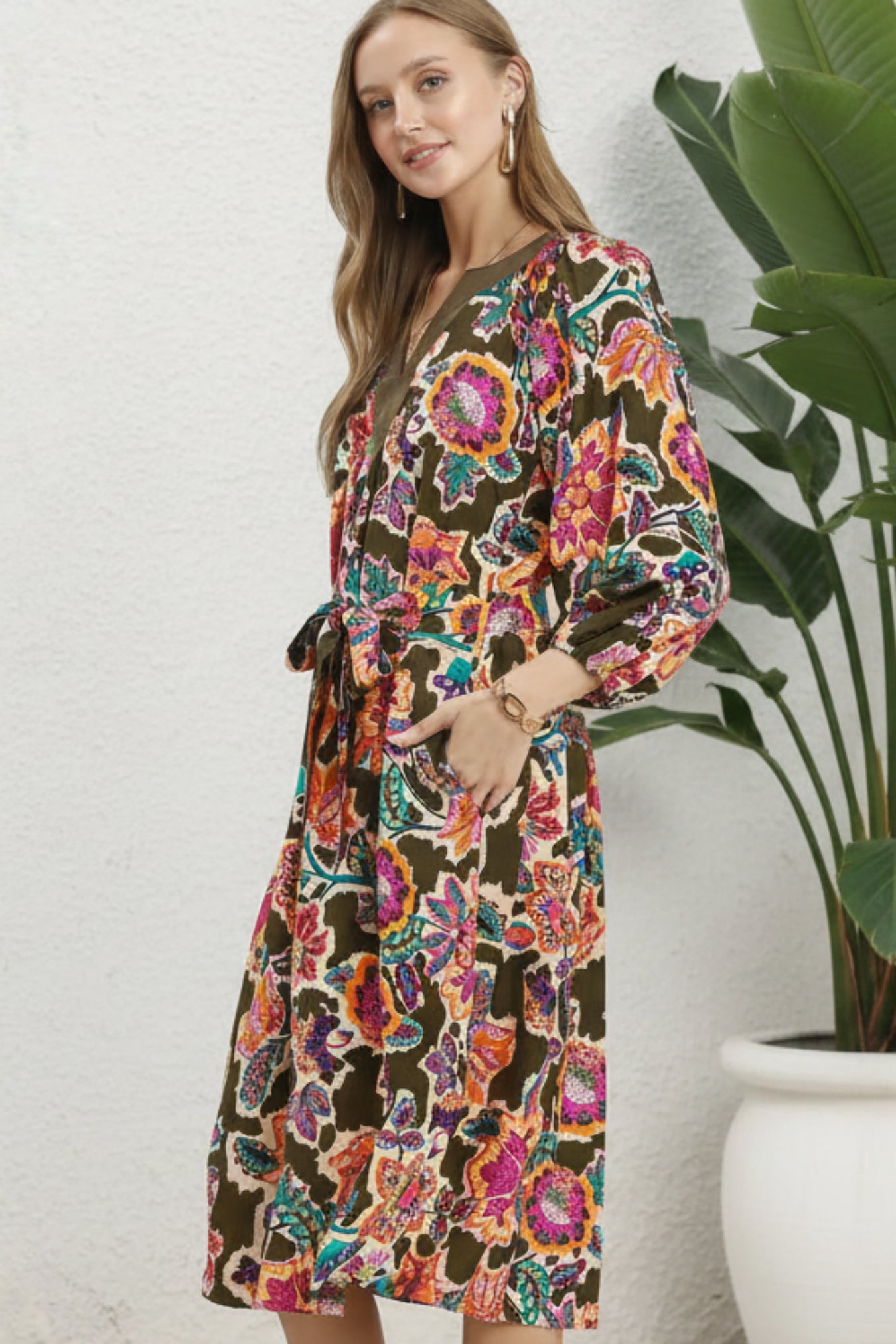A woman modeling a long-sleeve wrap dress with a vibrant multicolor floral print on an olive brown base, standing next to a potted plant against a textured white wall.