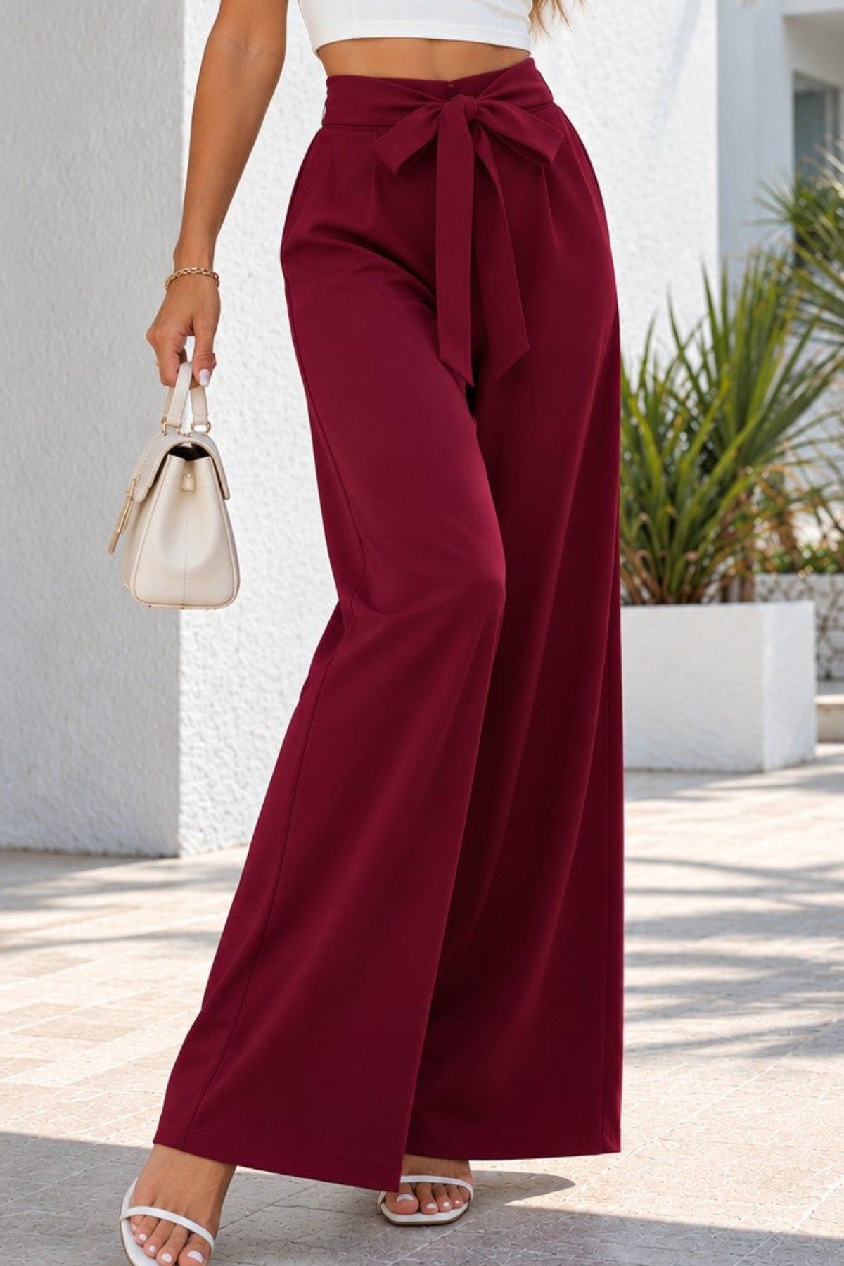 A woman wearing a white strapless crop top and high-waisted burgundy wide-leg pants with a front tie, walking outdoors in front of a white stucco building with palm trees.