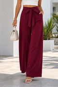 A woman wearing a white crop top and high-waisted burgundy wide-leg pants with a tie waist, walking outdoors in front of a white building with palm trees.