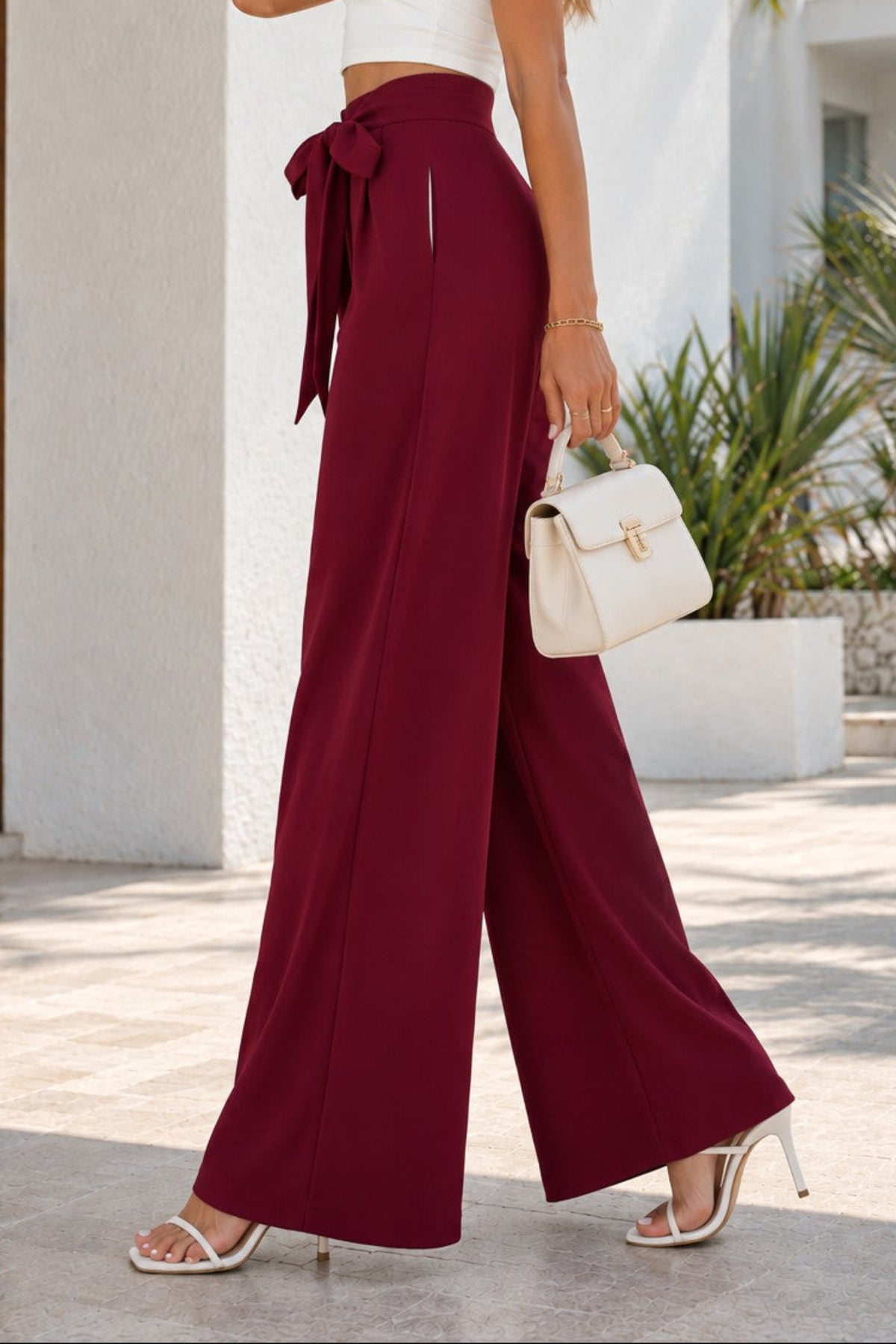 A woman wearing a white halter crop top and high-waisted burgundy wide-leg pants with a side tie, walking outdoors in front of a white building with palm trees.