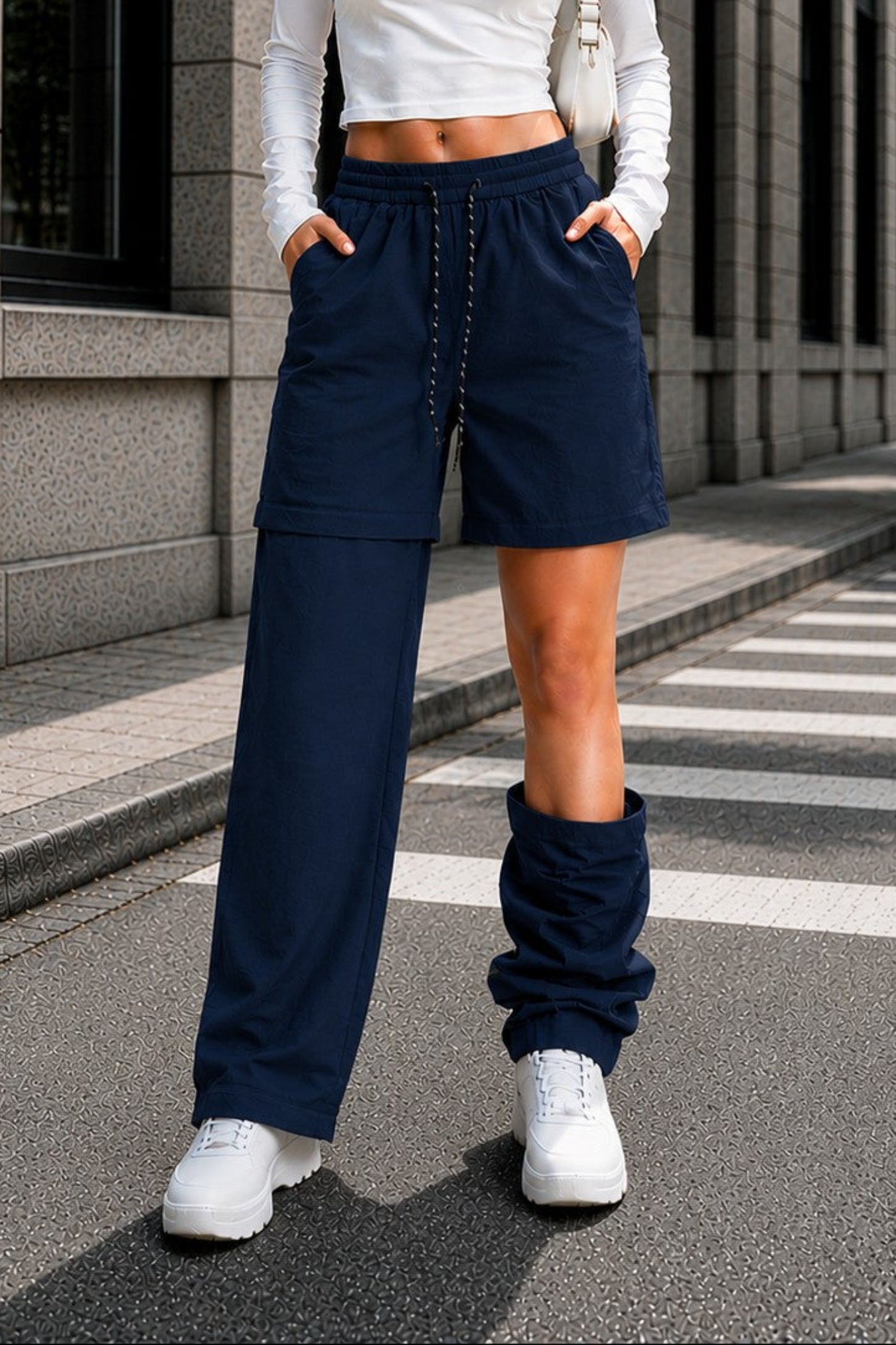 A woman stands on a city street wearing a white ribbed long-sleeve crop top, navy wide-leg shorts with drawstrings, matching navy leg warmers, and white sneakers.