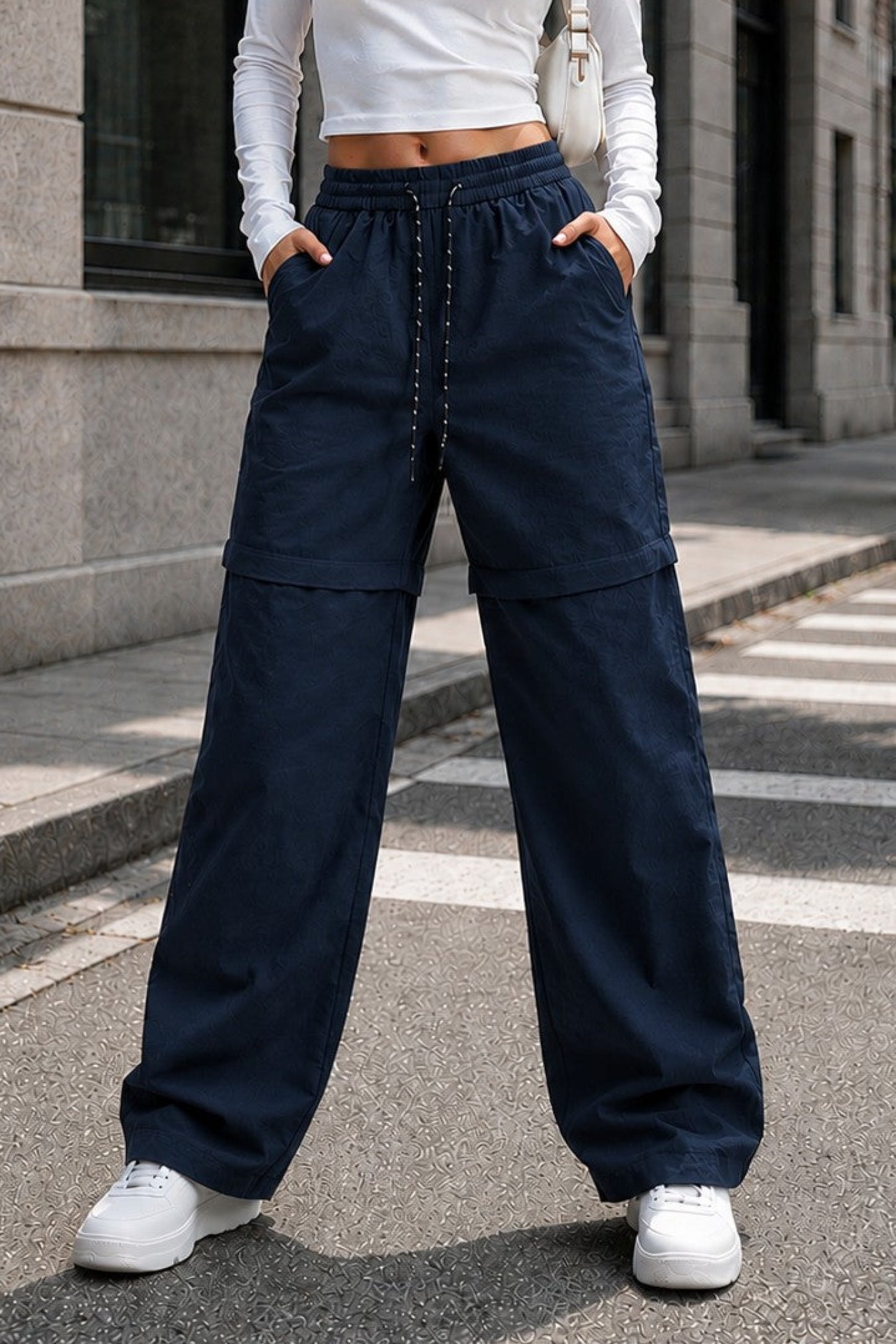 A woman stands on a city street wearing a fitted white long-sleeve crop top and dark navy wide-leg drawstring pants with white sneakers.
