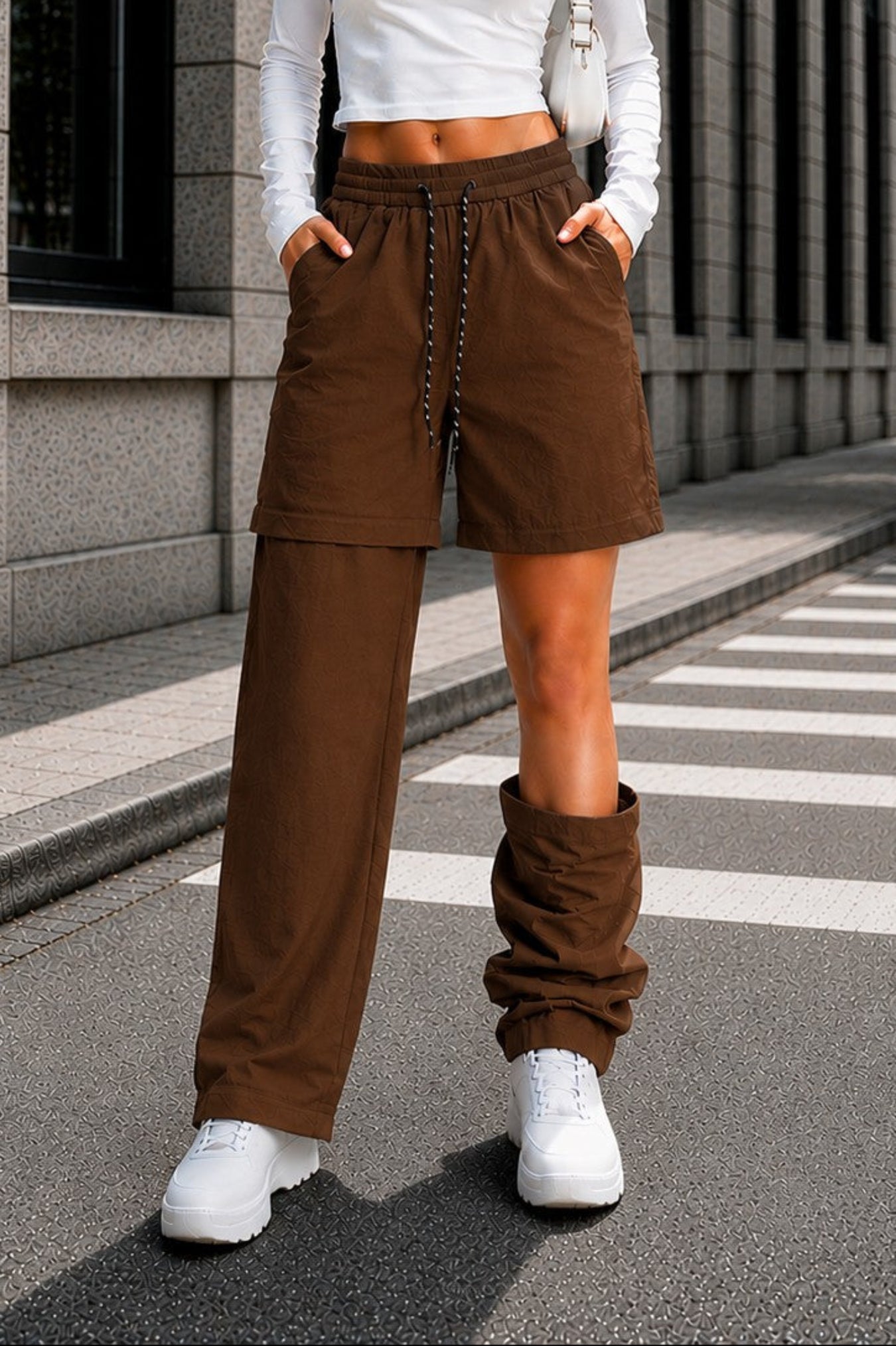 A woman stands on a city street wearing a white long-sleeve crop top, brown drawstring shorts, matching brown wide-leg pants, and white sneakers.