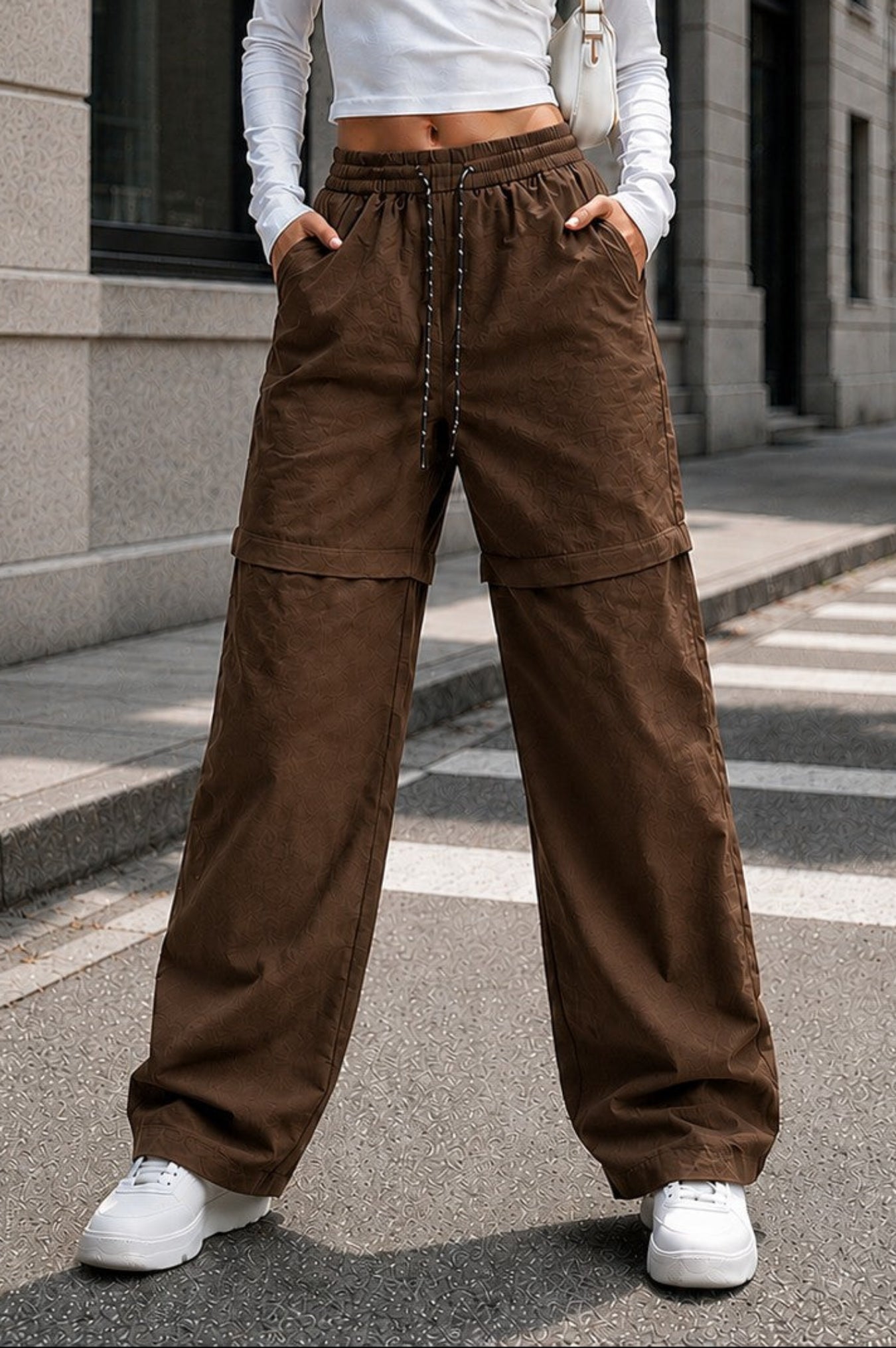 A woman stands on a city street wearing brown wide-leg cargo pants with a drawstring waist, a fitted white long-sleeve crop top, and white sneakers.