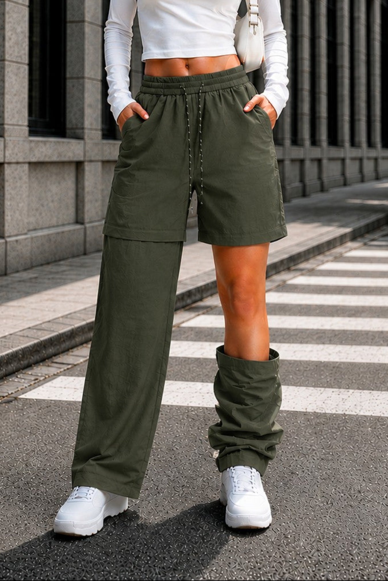 A woman stands on a city street crosswalk wearing a white long-sleeve crop top and olive green wide-leg shorts with matching green leg warmers, white sneakers, and a white shoulder bag.