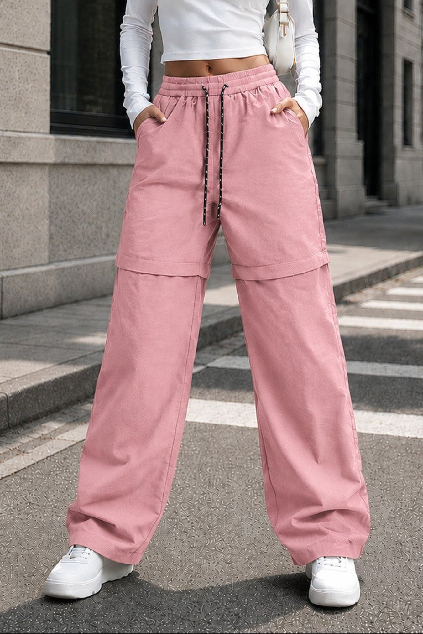 A woman stands on a city street wearing a white long-sleeve crop top and wide-leg pink drawstring pants with white sneakers.