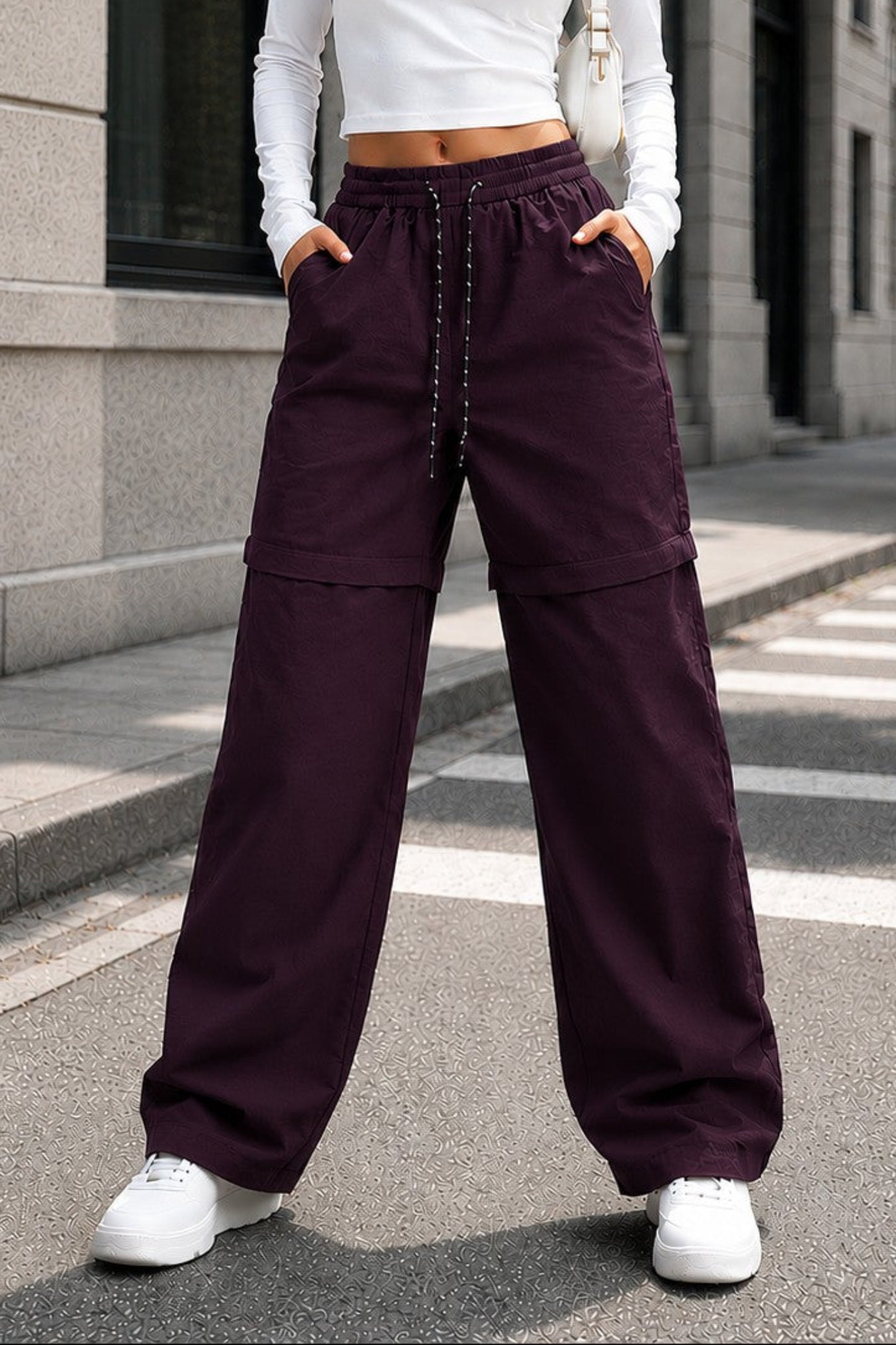 A woman stands on a city street wearing a white long-sleeve crop top and dark plum wide-leg drawstring pants with white sneakers.