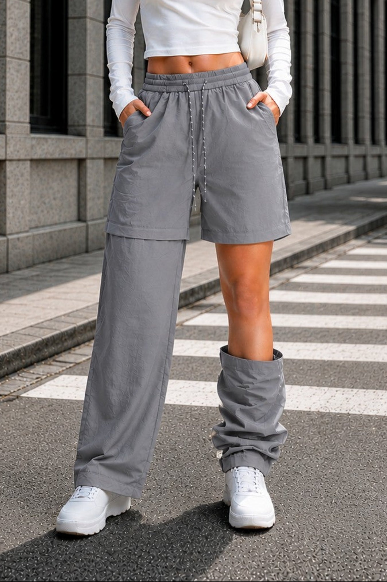 A woman stands on a city crosswalk wearing a white long-sleeve crop top, light gray drawstring shorts, and matching gray leg warmers with white sneakers. She is posed in front of a modern gray stone building with trees on the sidewalk.