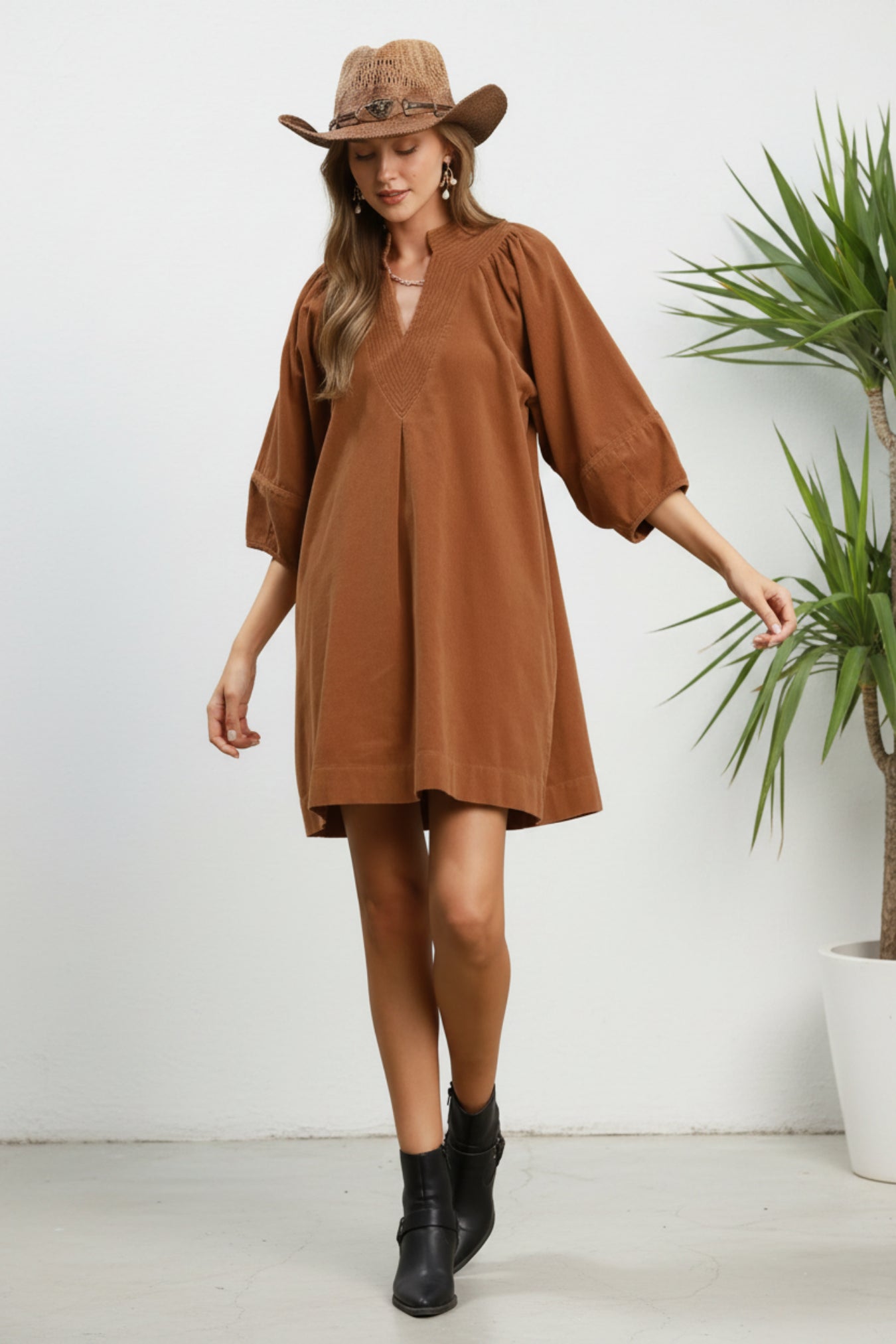 Woman wearing a brown dress and hat on a white background next to a plant