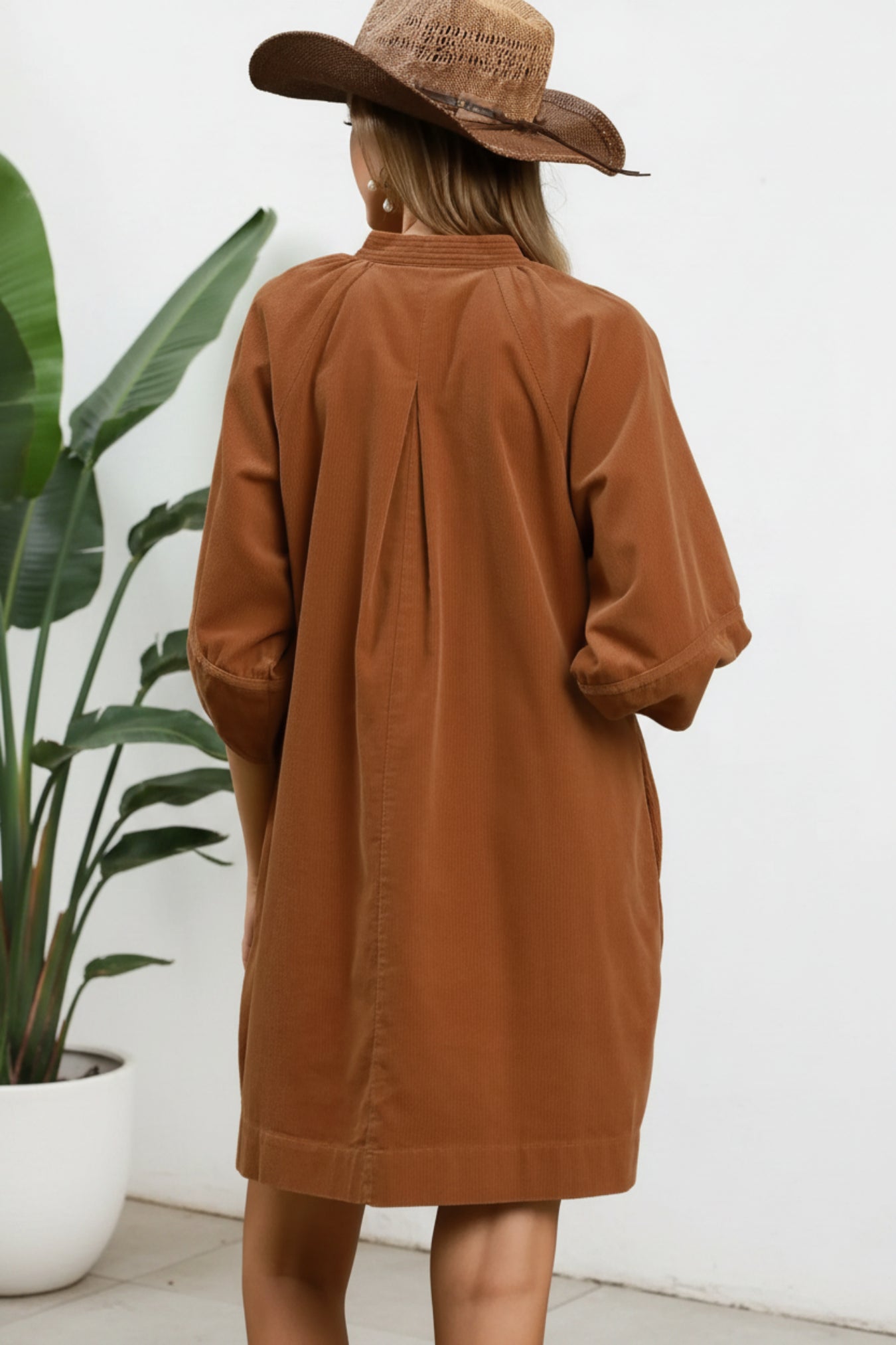 A woman shown from the back wearing a camel-brown corduroy shirt dress and a straw cowboy hat against a white background.