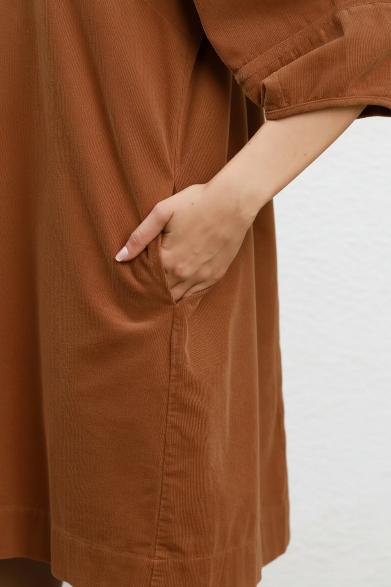 Close-up of a person wearing a camel brown corduroy dress, showing the side pocket with a hand inserted, against a white background.