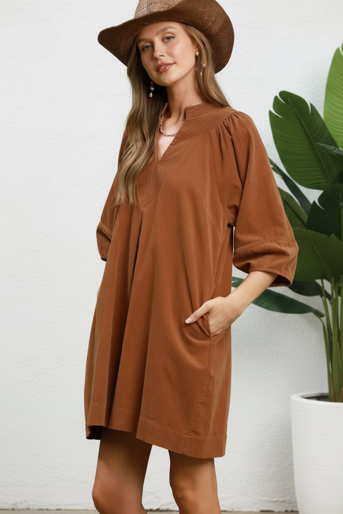 A woman wearing a camel brown short-sleeve tunic dress with a V-neck and side pockets, paired with a matching brown hat, against a white background next to a plant.