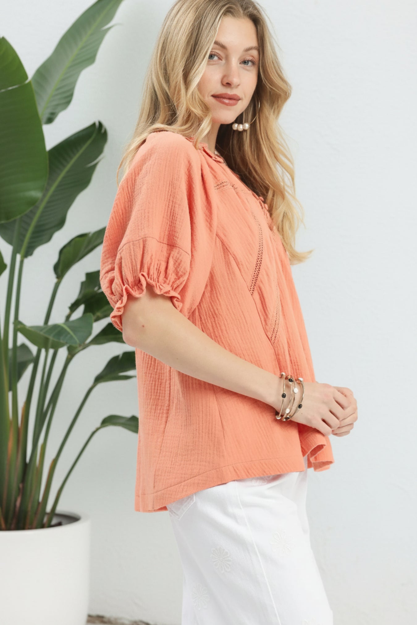 Woman wearing a peach blouse standing next to a plant against a white wall