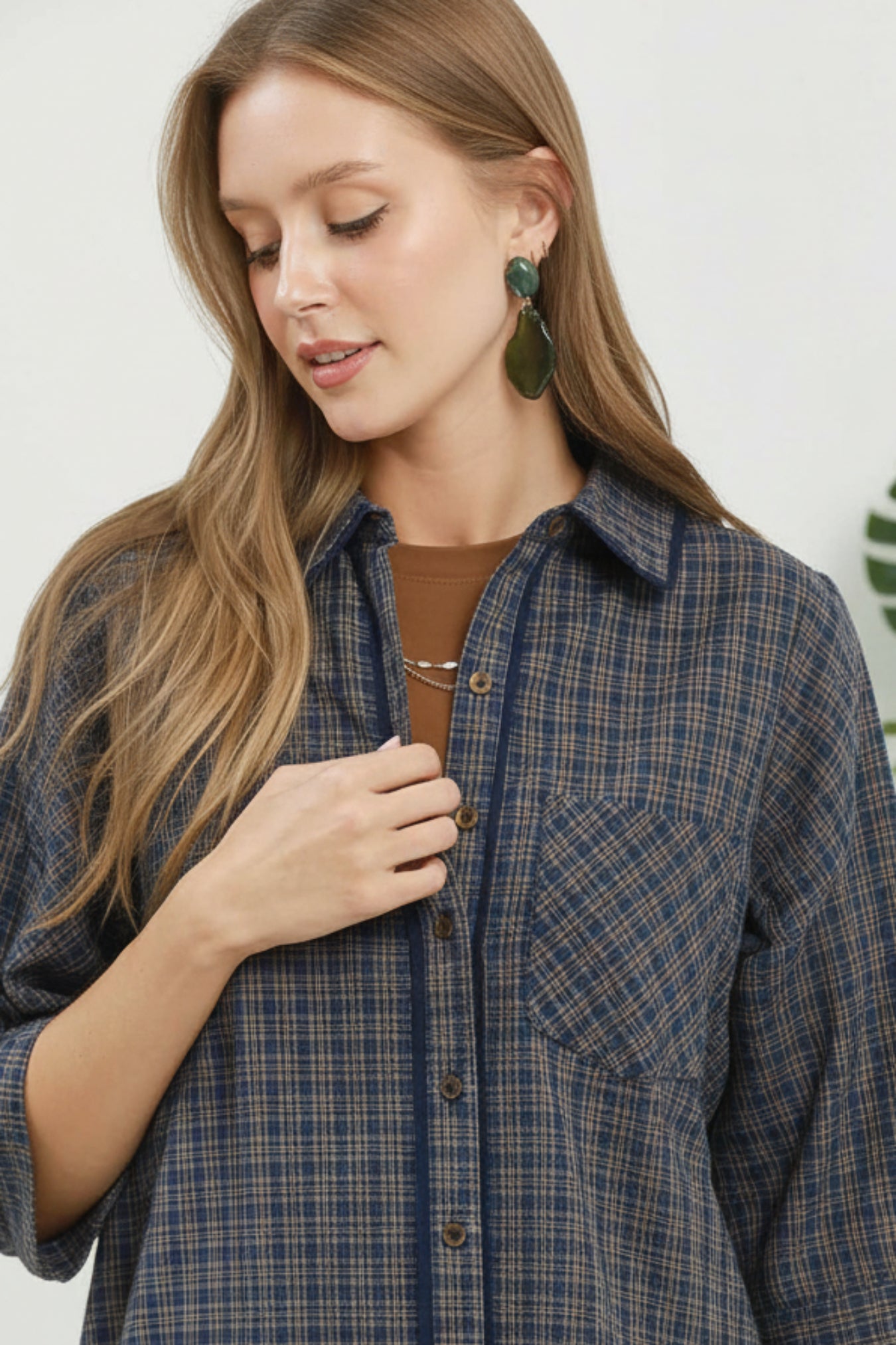 A woman wearing a navy blue and brown plaid button-down shirt, styled with a brown top underneath and green teardrop earrings, shown in a close-up shot against a light background.
