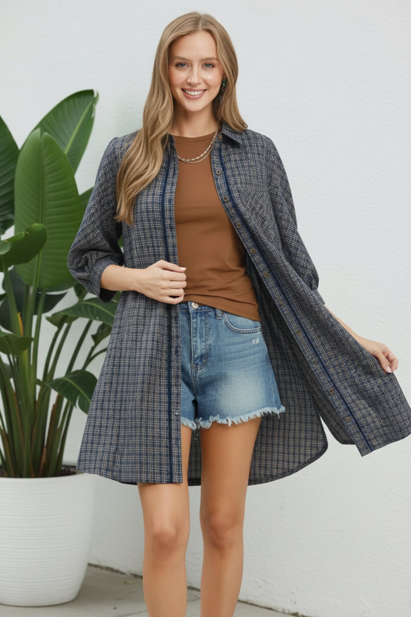 Woman wearing a navy plaid shirt over a brown top and denim shorts on a white background with a plant