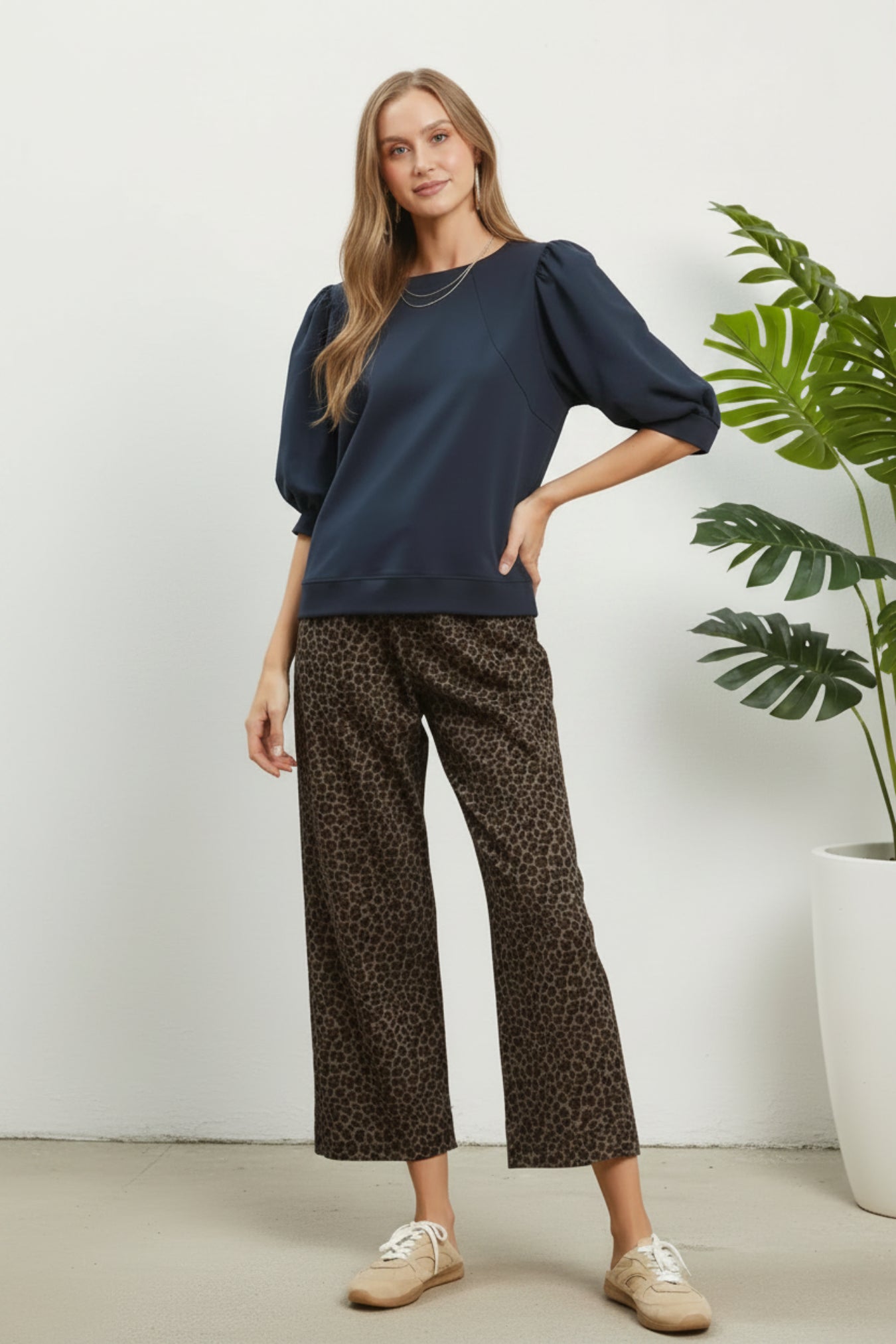 A woman modeling a navy blue long-sleeve top with puffed 3/4 sleeves and dark brown leopard-print cropped pants, standing against a white background