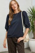 A woman modeling a navy blue long-sleeve top with puff sleeves, paired with leopard-print pants and a cream crossbody bag, against a white background.