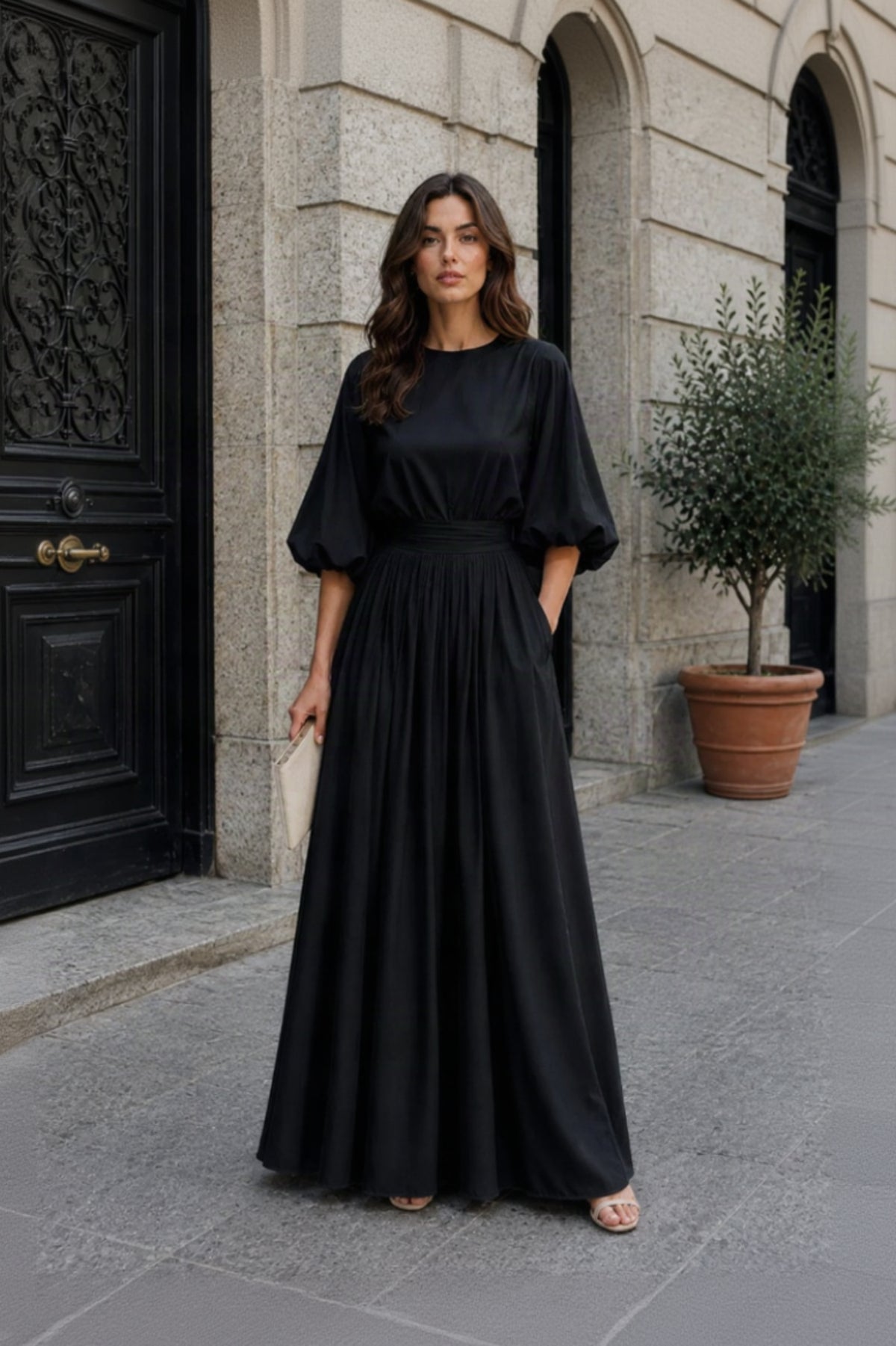 A woman stands outdoors in front of a stone building, wearing a long black maxi dress with voluminous elbow-length puff sleeves, a fitted waist, and side pockets. She is holding a small light beige clutch and wearing flat sandals.