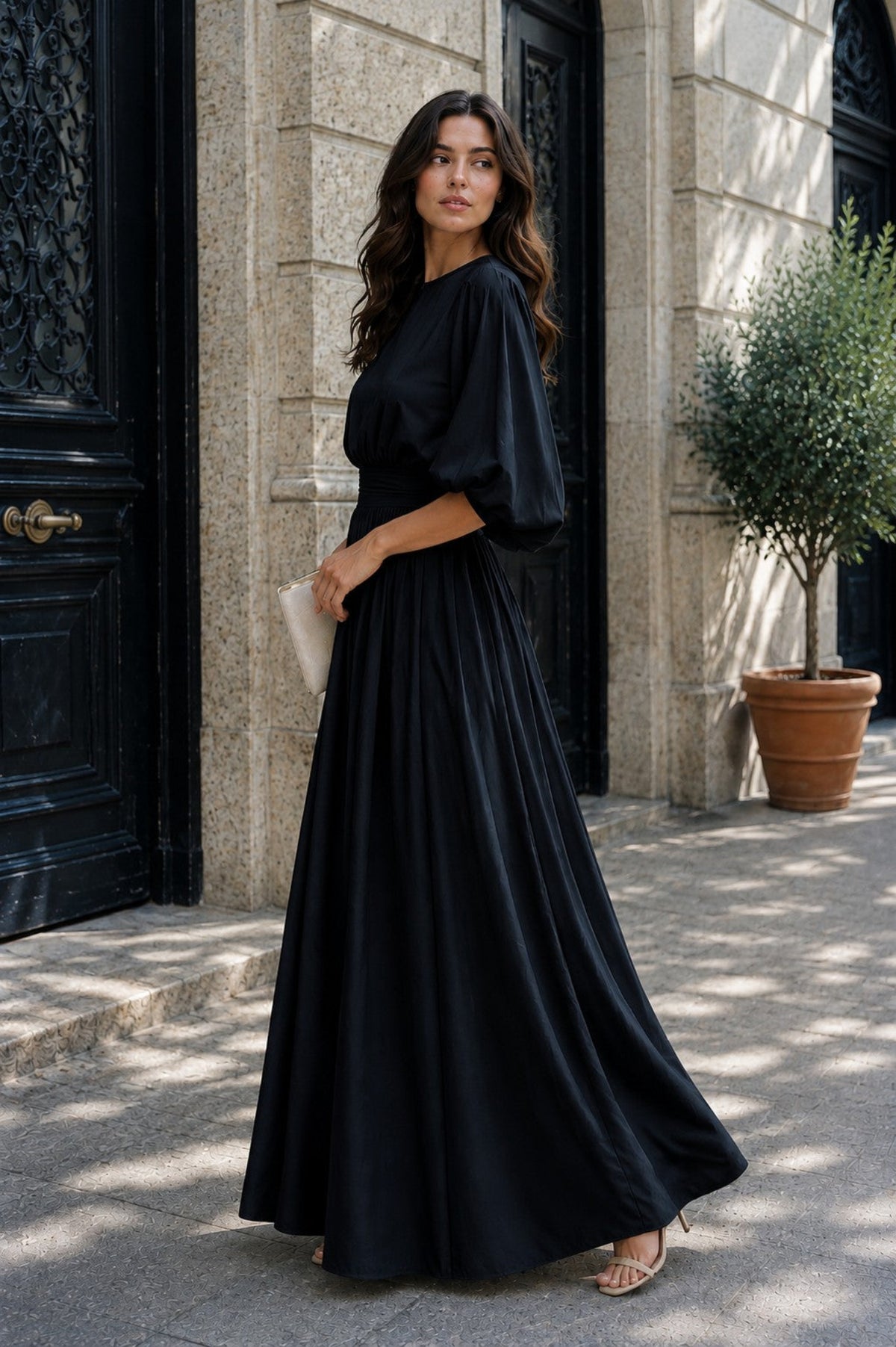 A woman wearing a flowing black maxi dress with voluminous short sleeves stands on a stone sidewalk in front of a building entrance, holding a small white clutch.