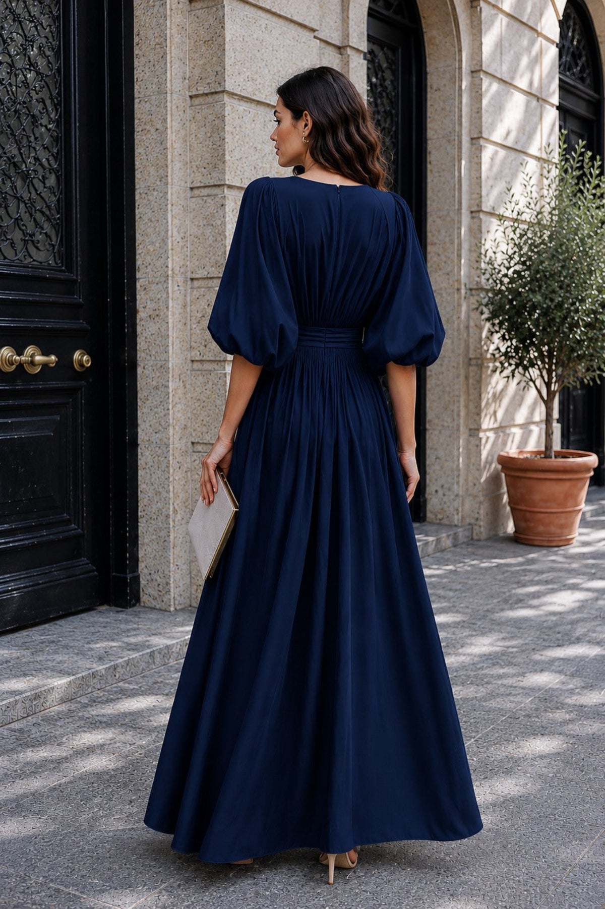 A woman wearing a navy blue maxi dress with puffed elbow-length sleeves and a defined waist, photographed from the back in an outdoor setting in front of a stone building.
