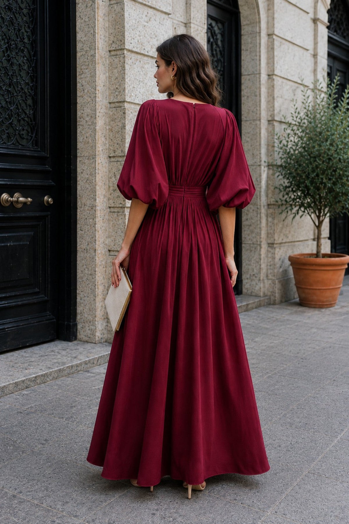 A woman wearing a long burgundy maxi dress with puffed elbow-length sleeves and a gathered waist, standing outdoors on a stone walkway in front of a building with a black door and potted plant.