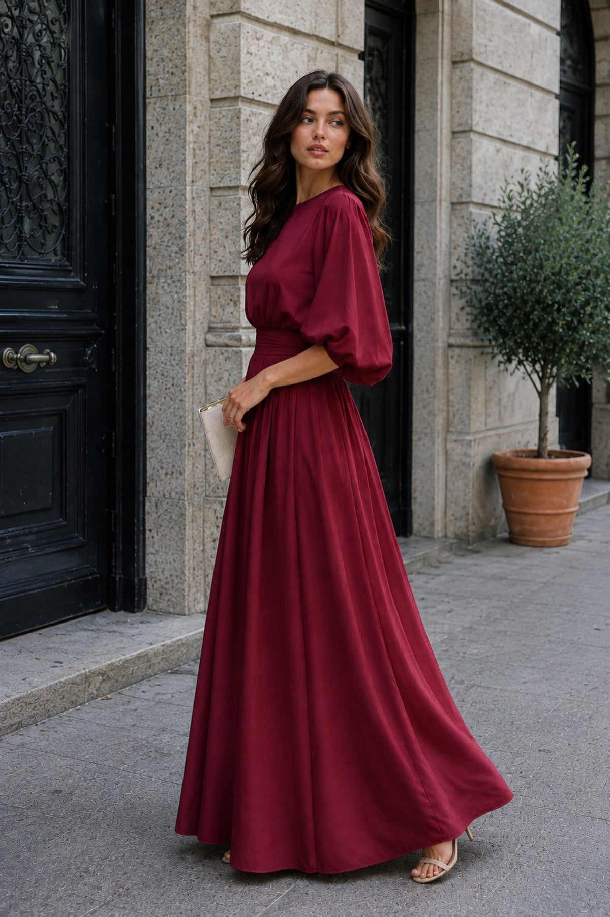 A woman wearing a long, flowing burgundy maxi dress with voluminous puff sleeves stands on a city sidewalk in front of a stone building with black doors.