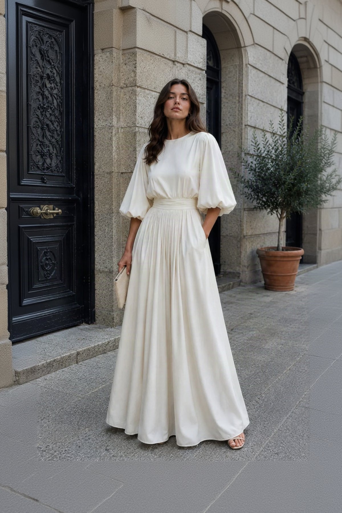 A woman stands on a stone sidewalk wearing a long cream maxi dress with voluminous elbow-length puff sleeves, a fitted waist, and a flowing skirt. She holds a small cream clutch and wears flat sandals.