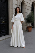 A woman stands on a city sidewalk in front of a stone building, wearing a long cream maxi dress with voluminous elbow-length puff sleeves, a defined waist, and a flowing skirt. She holds a small cream clutch.