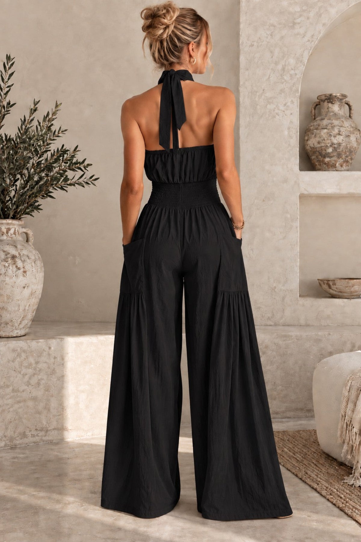 A woman viewed from behind wearing a black halter-neck wide-leg jumpsuit in a rustic beige interior with stone walls, a plant, and a textured vase.