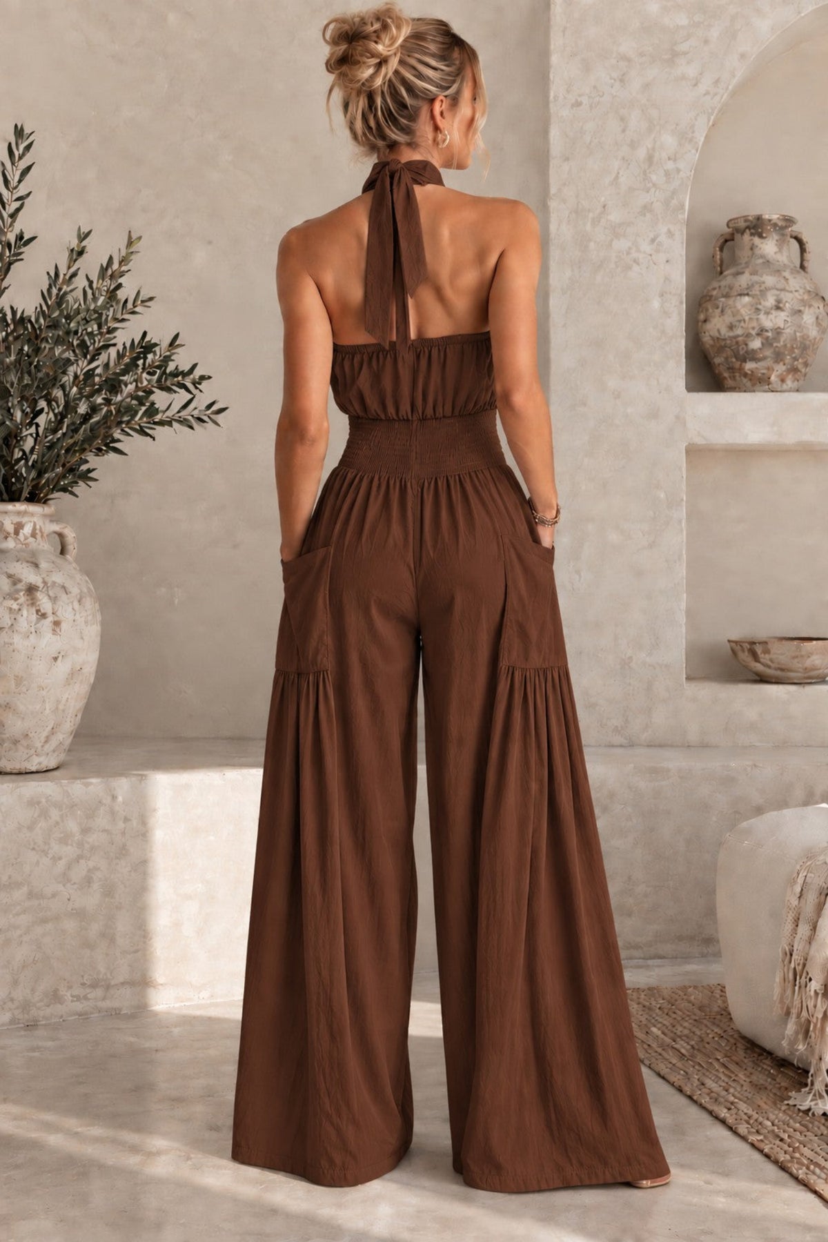 A woman wearing a brown halter-neck wide-leg jumpsuit, shown from the back in a rustic, sunlit room with textured walls and stone shelves.