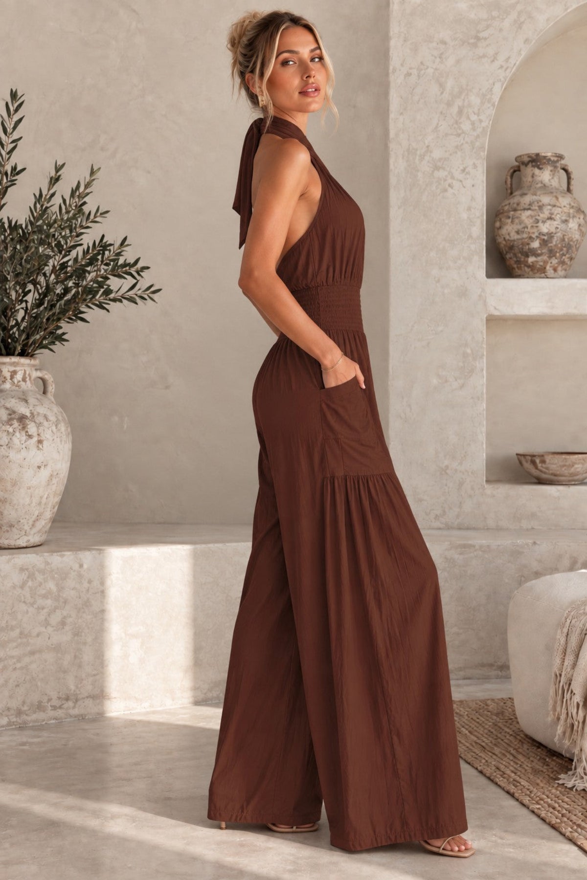 A woman wearing a brown halter-neck wide-leg jumpsuit with side pockets, standing in a sunlit, rustic interior with textured walls, a plant, and a vase.