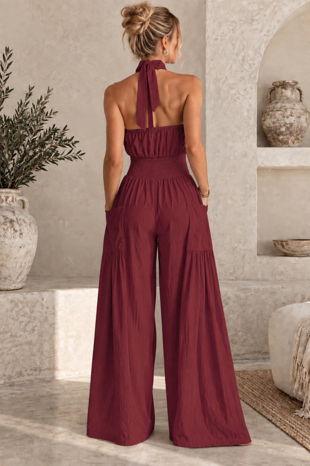A woman viewed from behind wearing a sleeveless burgundy wide-leg jumpsuit with a tie-neck detail, standing in a rustic beige interior with stone walls and decorative vases.