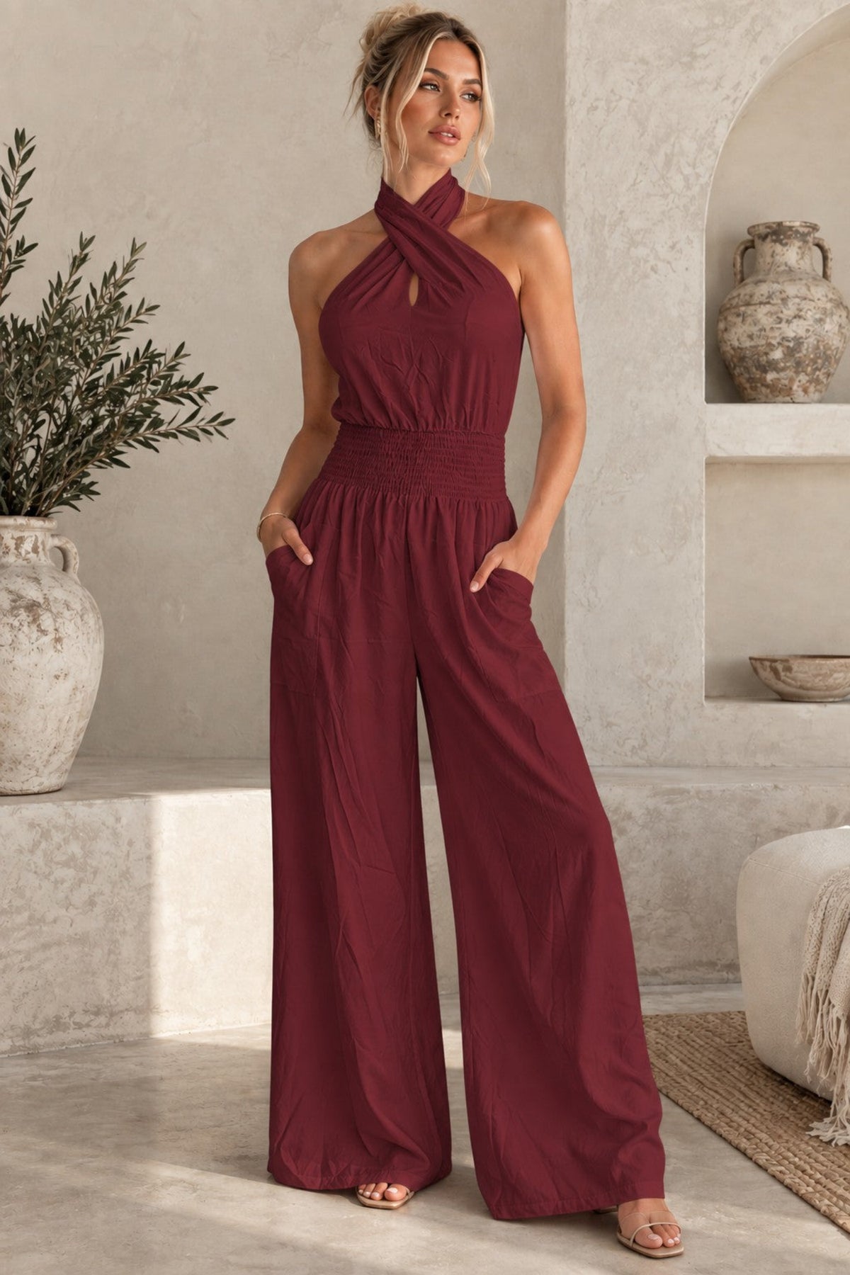 A woman models a maroon halter-neck jumpsuit with a keyhole cutout at the collar and wide legs, standing in a rustic, beige-walled room with a textured stone floor.