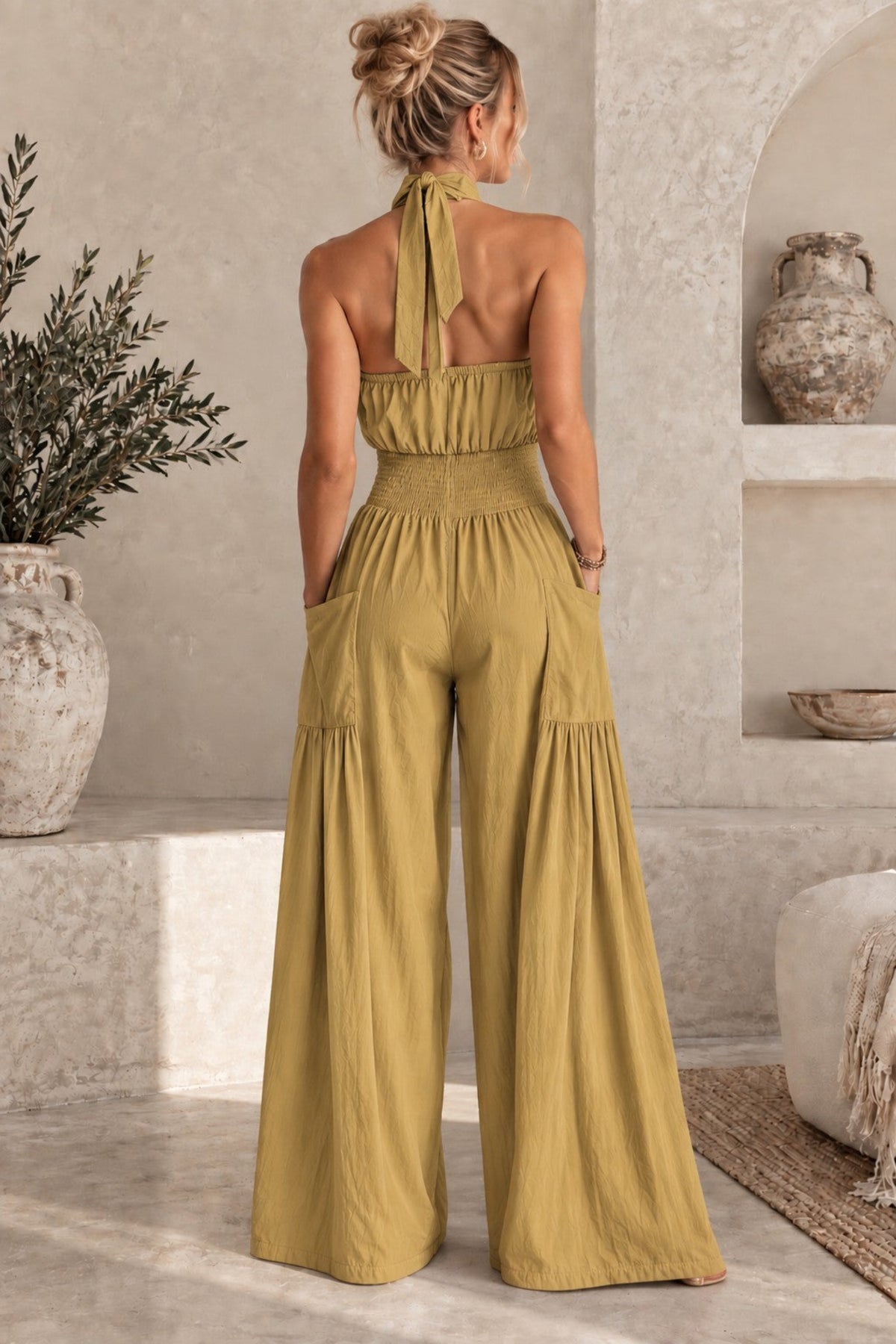 A woman stands with her back to the camera wearing a mustard yellow halter-neck wide-leg jumpsuit in a rustic, neutral-toned indoor setting.
