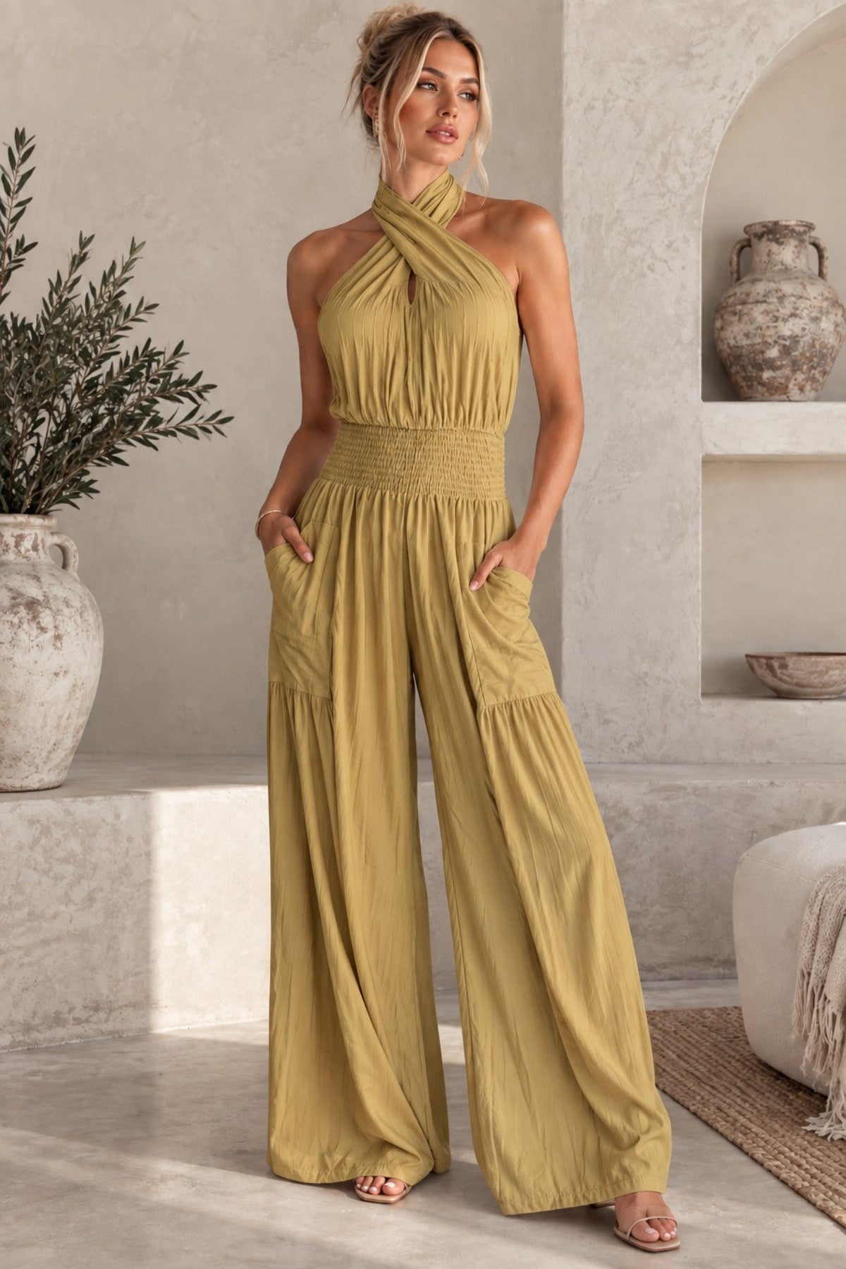 A woman models a mustard yellow halter-neck wide-leg jumpsuit in a sunlit, rustic interior setting with textured walls, a plant, and a shelf.