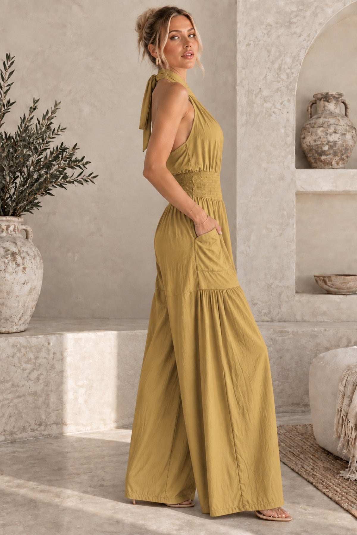 A woman modeling a mustard yellow halter-neck maxi jumpsuit with wide legs and side pockets, standing in a rustic, sunlit stone room with textured walls and decorative pottery.