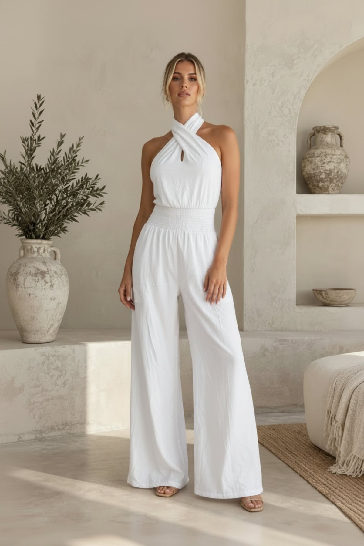 A woman models a white halter-neck wide-leg jumpsuit in a sunlit, rustic beige interior with textured walls, a plant, and a ceramic vase.