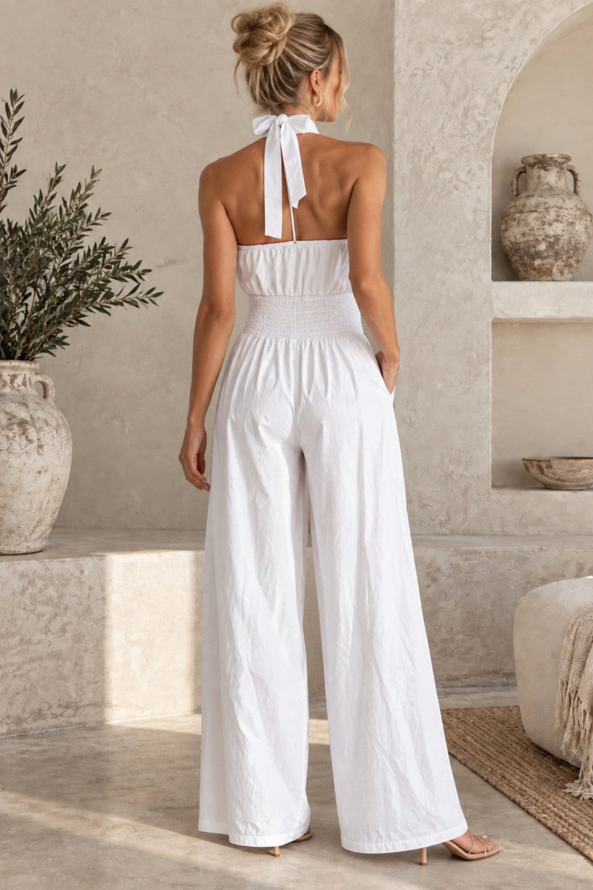 A woman viewed from behind wearing a white halter-neck wide-leg jumpsuit in a sunlit neutral interior with textured walls, a plant, and a vase.