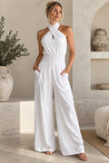 A woman modeling a white halter-neck wide-leg jumpsuit in a sunlit, rustic beige interior with textured walls, a shelf, and large ceramic vases.