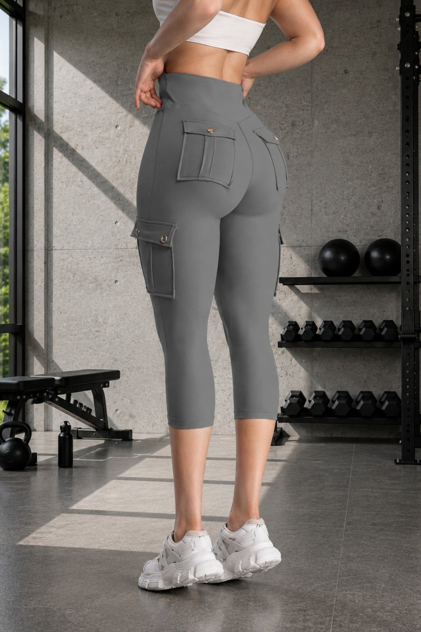 A woman in a gym wearing high-waisted gray cargo capri leggings, shown from the back. The leggings feature large side cargo pockets and a high-rise waist, paired with white sneakers.