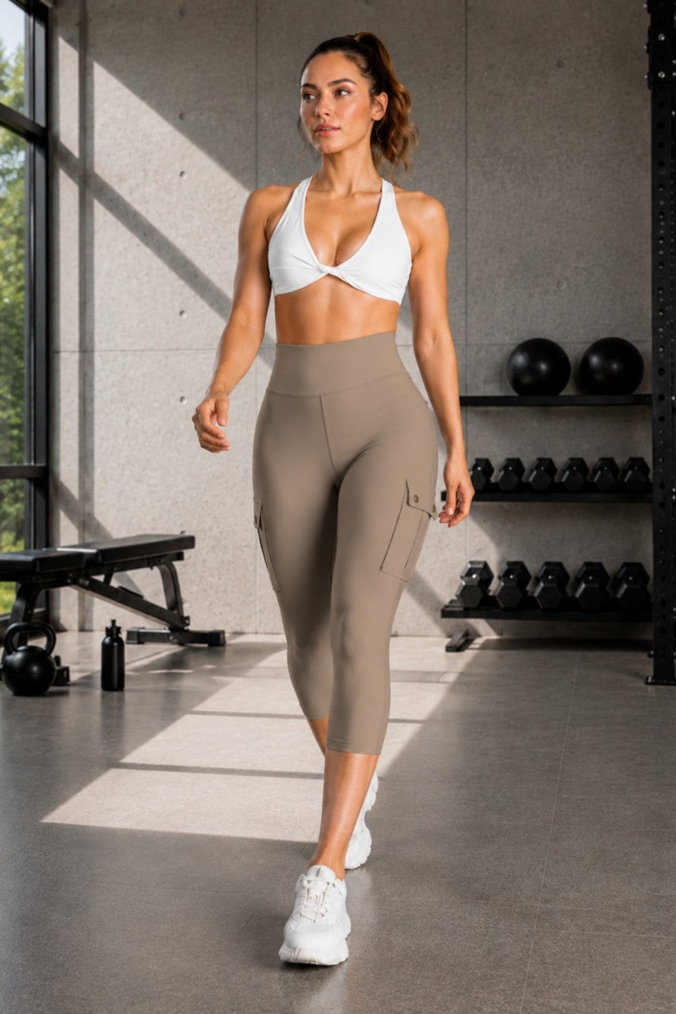 A woman walks through a modern gym wearing a matching two-piece activewear set consisting of a white sports bra and high-waisted taupe capri leggings with side cargo pockets.