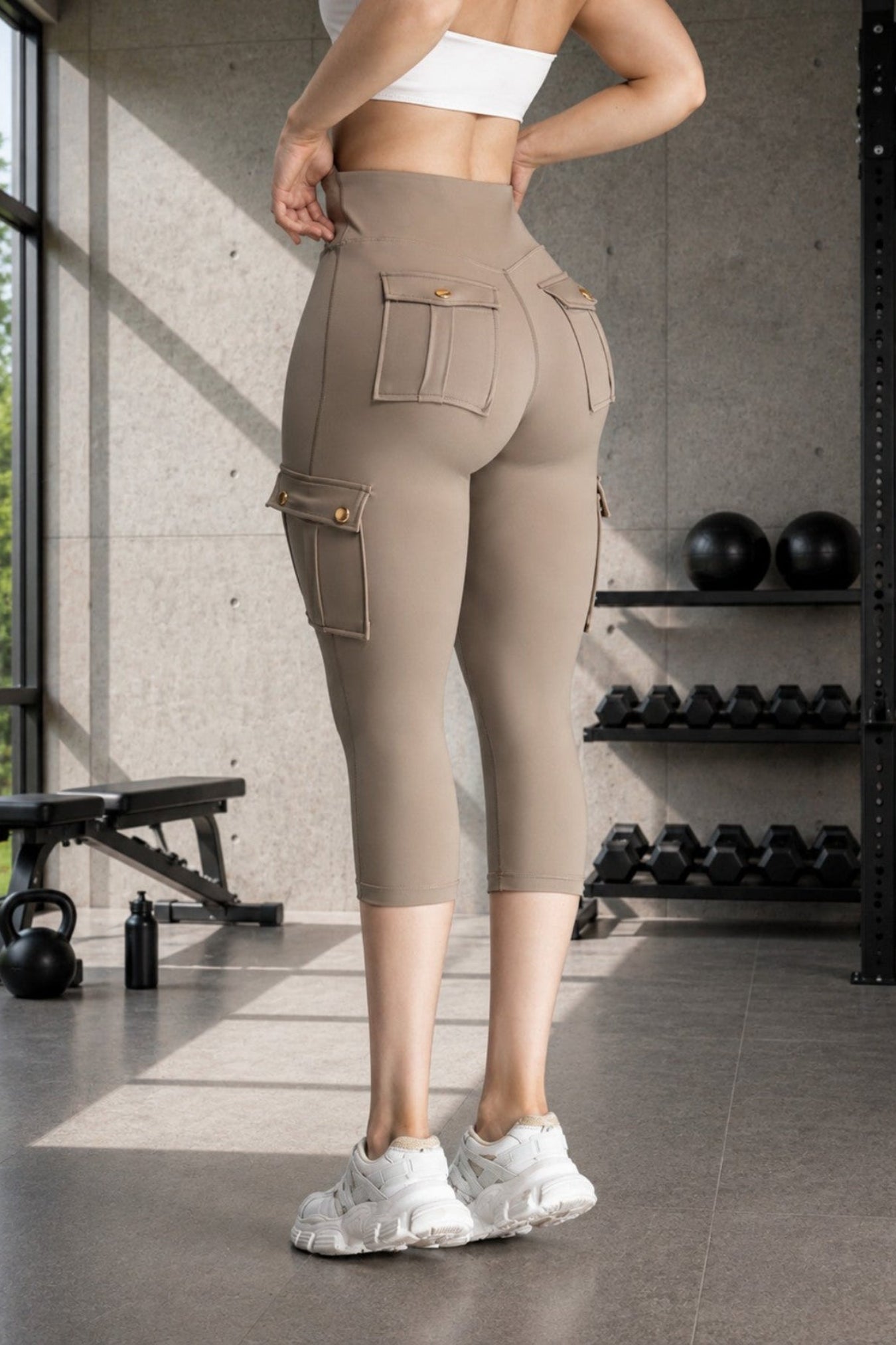A woman in a gym wearing high-waisted taupe cargo capri leggings, shown from the back. The leggings feature large side cargo pockets and a white sports bra, paired with white sneakers.