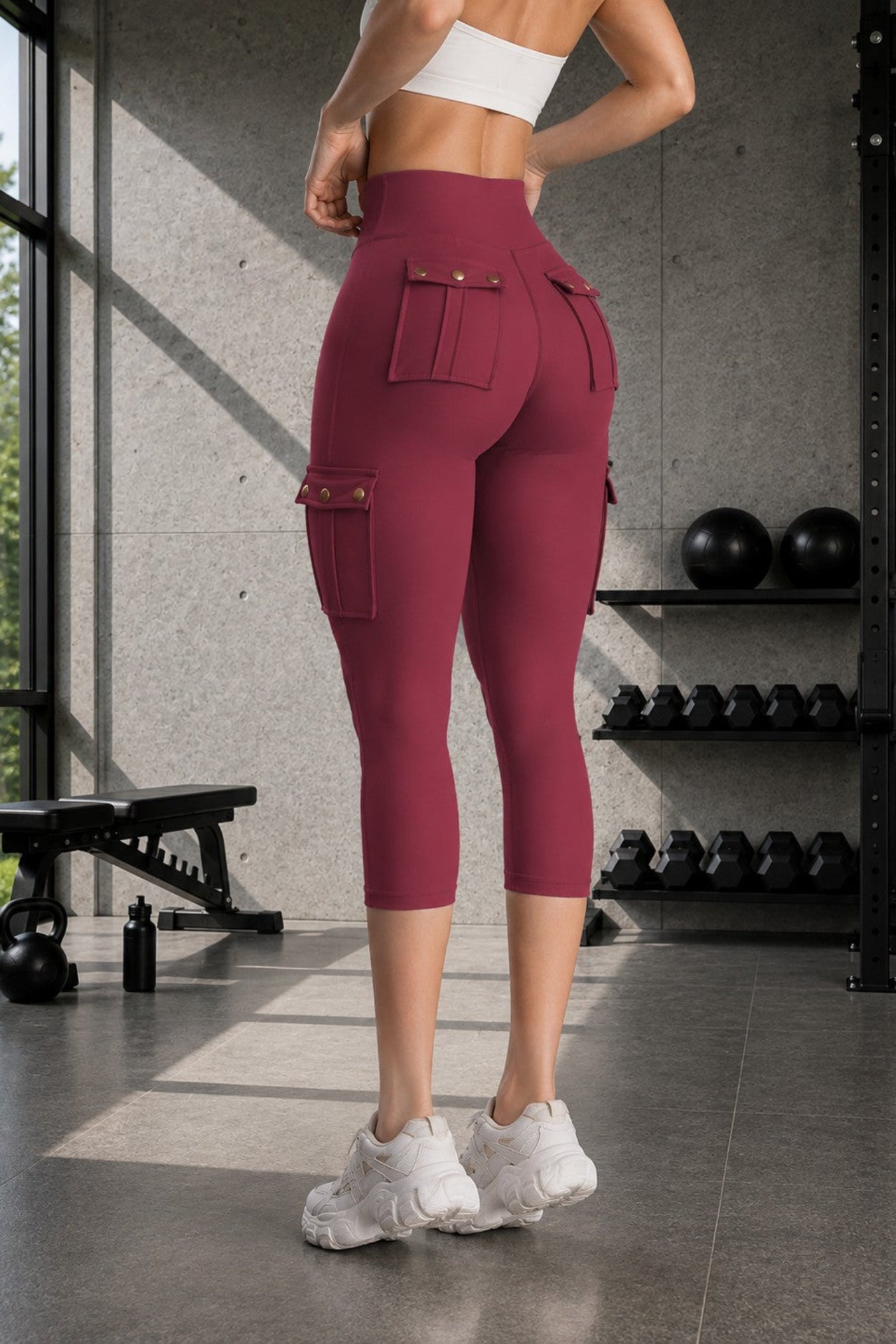 A woman in a gym wearing high-waisted burgundy cargo capri leggings, shown from the back. The leggings feature side cargo pockets and a white sports bra, with white sneakers on a concrete gym floor.