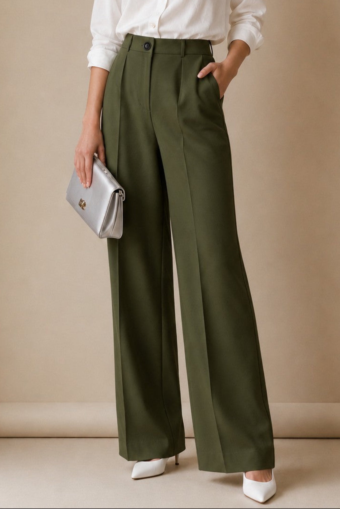 A woman stands against a warm beige studio backdrop wearing a white button-up blouse, olive green wide-leg trousers, and white pointed shoes, while holding a small metallic clutch.