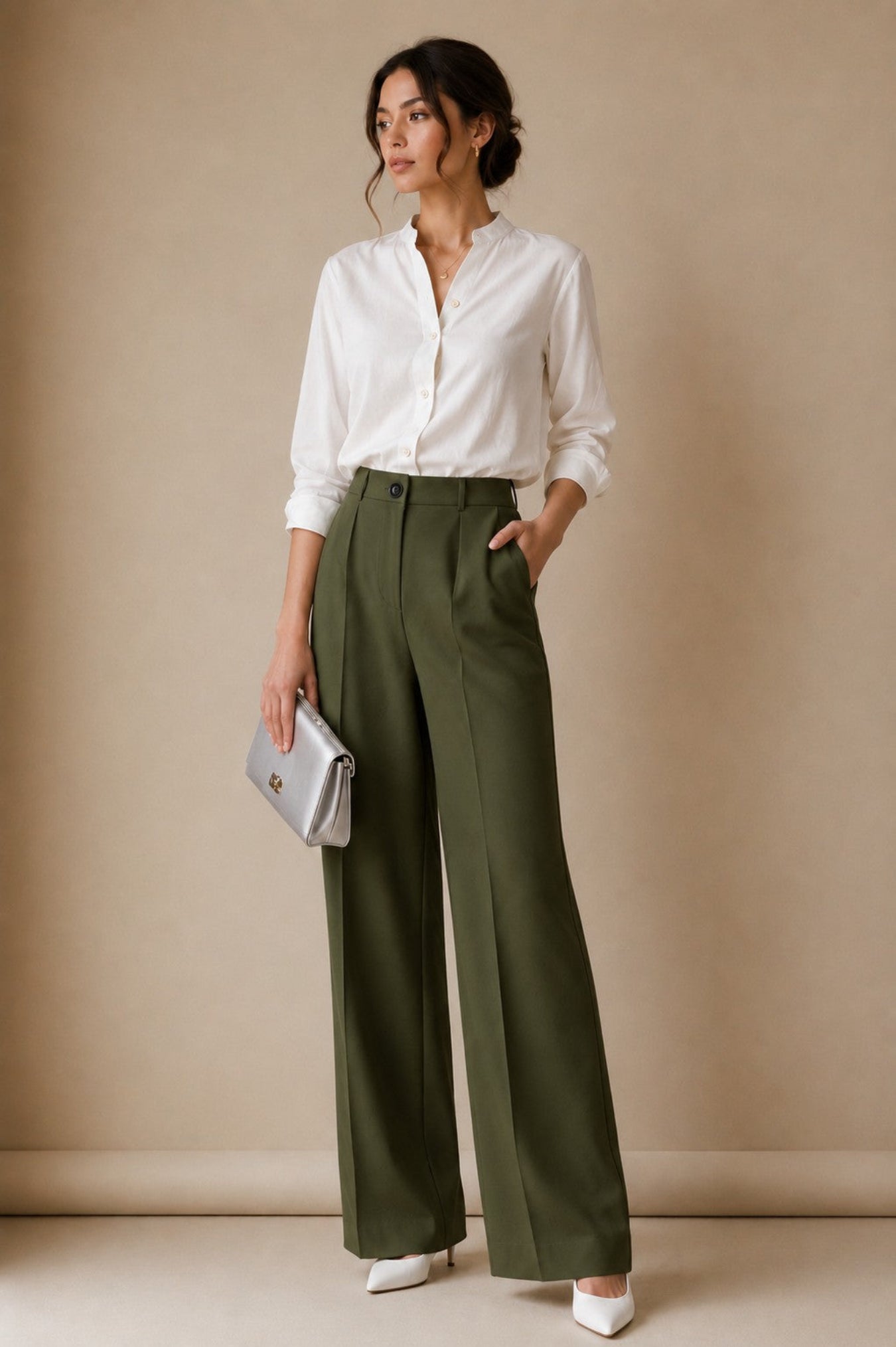 A woman stands against a warm beige studio backdrop wearing a white button-up blouse, olive green wide-leg trousers, and white pointed shoes, while holding a small metallic clutch.