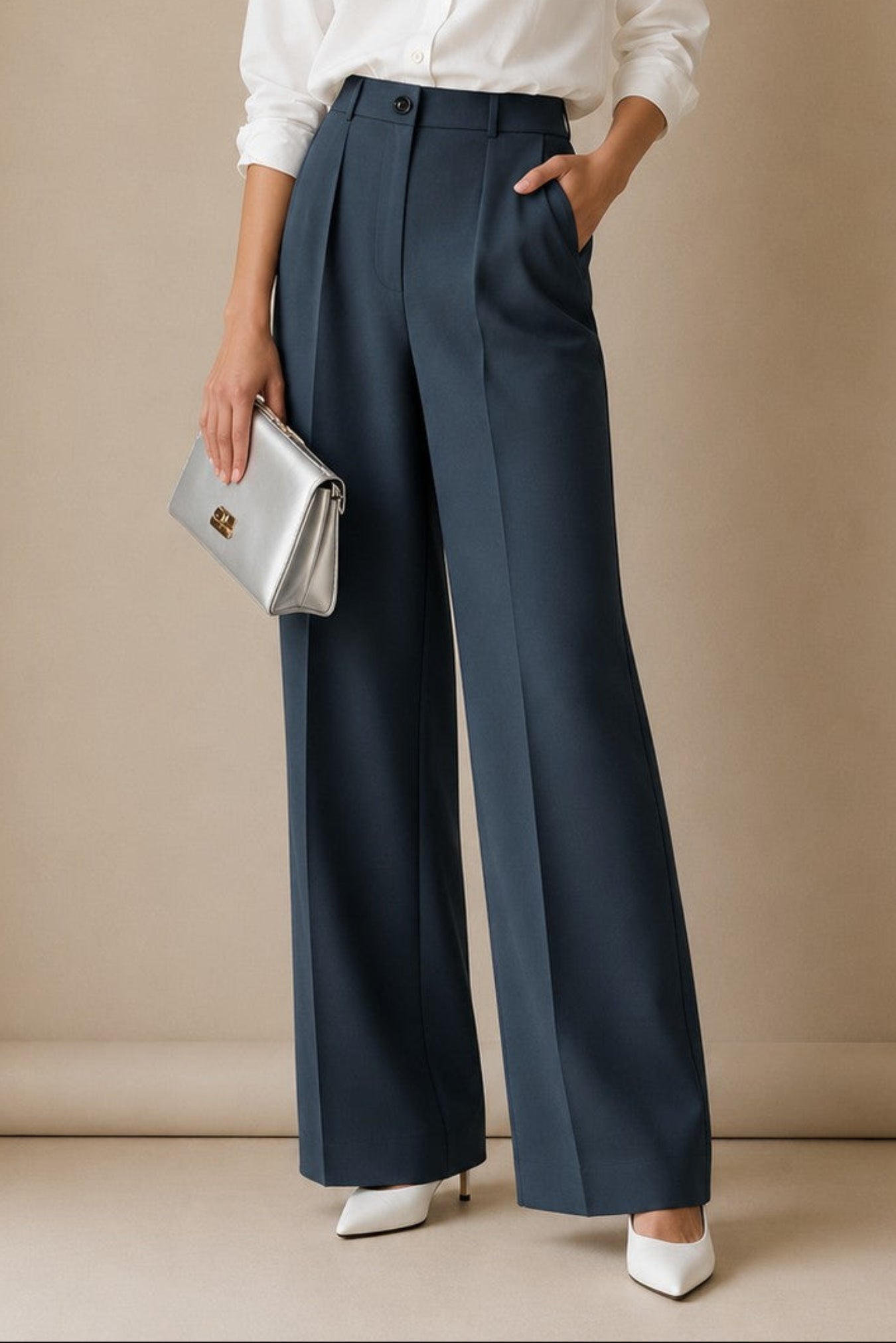 A woman stands against a beige studio backdrop wearing a white button-up blouse, navy wide-leg trousers, white pointed shoes, and holding a silver clutch.