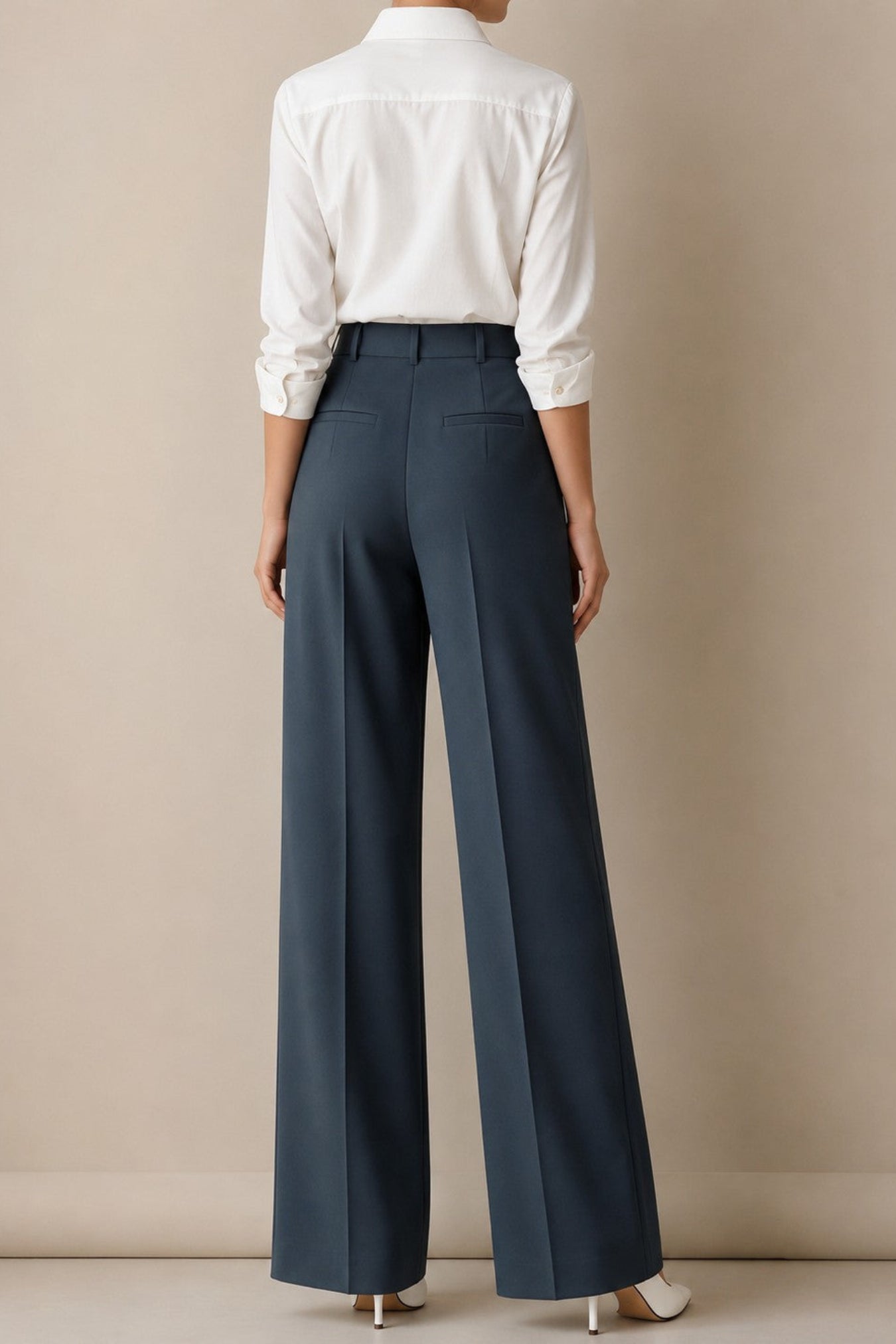 A model shown from the back wearing high-waisted, wide-leg navy trousers paired with a white button-down shirt and white heels, set against a plain beige studio background.