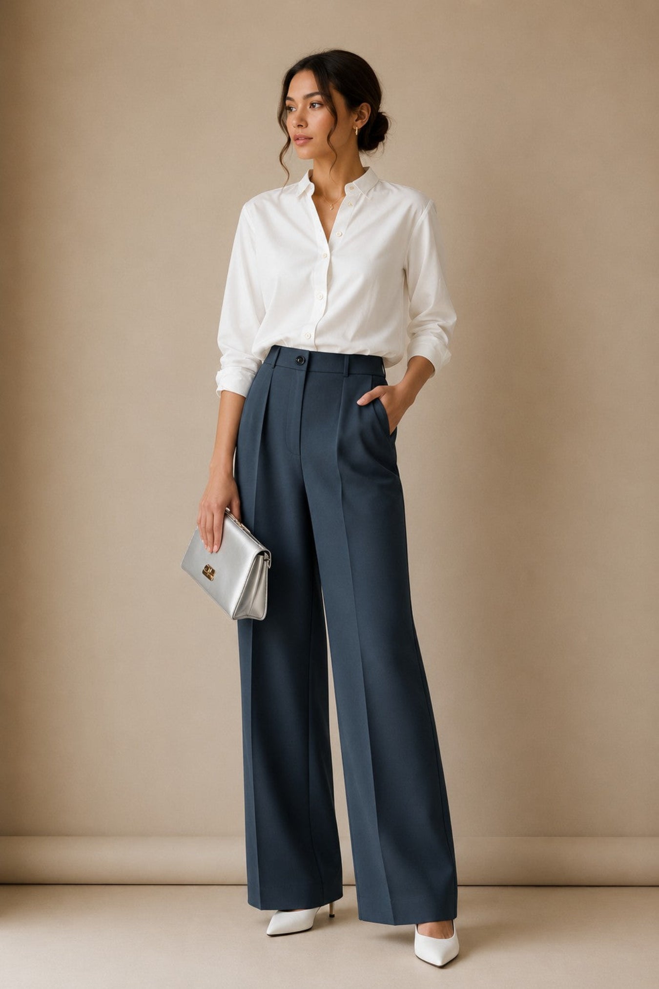 A woman stands against a beige studio backdrop wearing a white button-up blouse, navy wide-leg trousers, white pointed shoes, and holding a silver clutch.