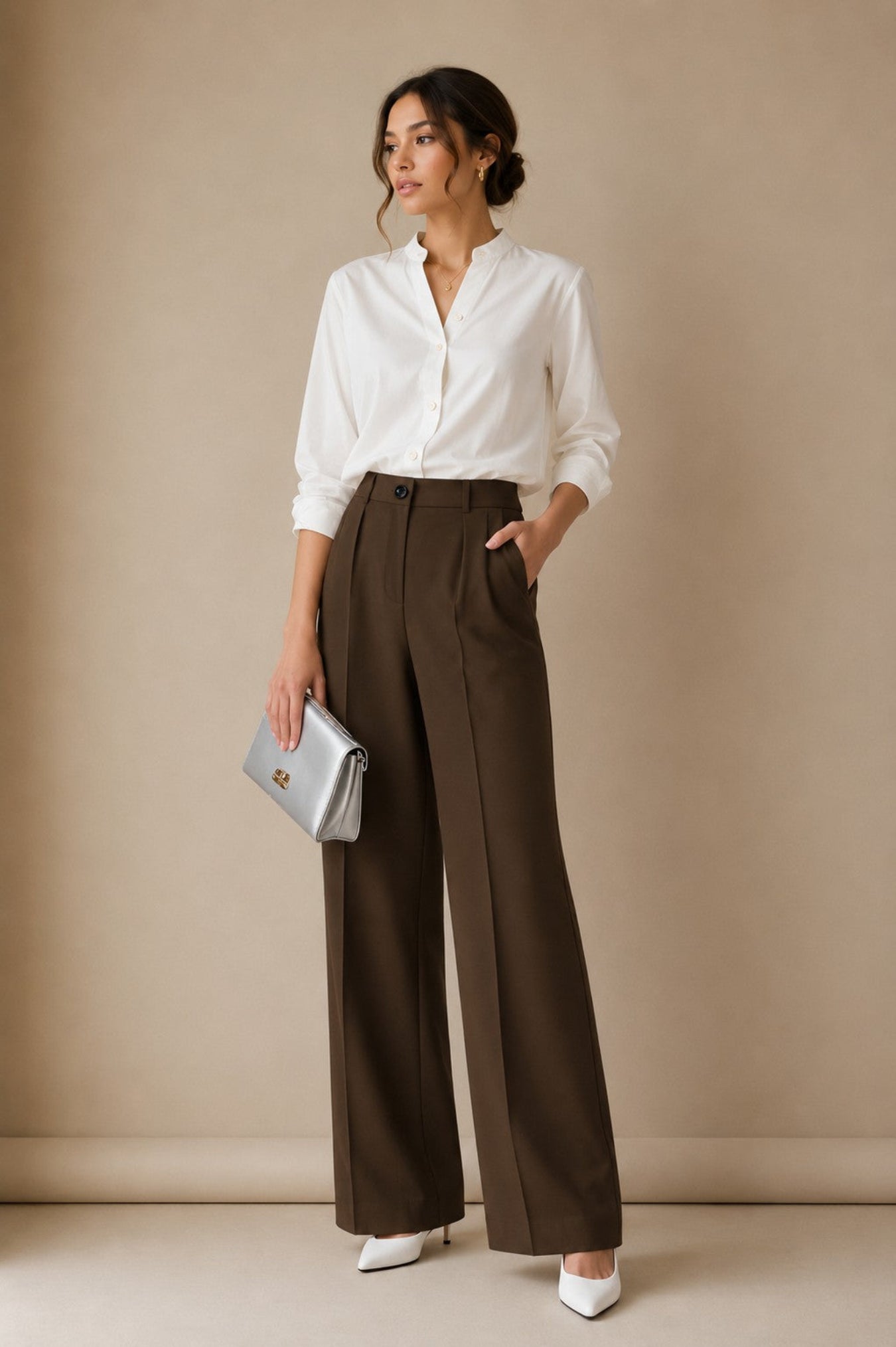 A woman stands in a studio wearing a white button-up blouse, dark brown wide-leg trousers, white pointed shoes, and holding a silver clutch.