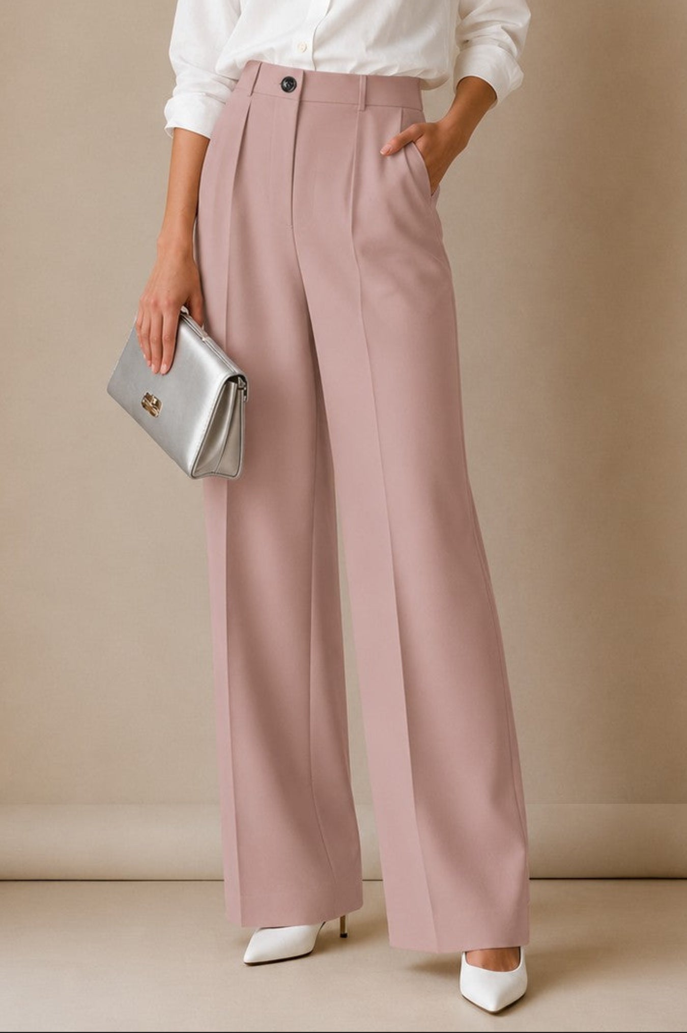 A woman stands in a studio wearing a white button-up shirt, dusty pink wide-leg trousers, white pointed shoes, and holding a silver clutch.